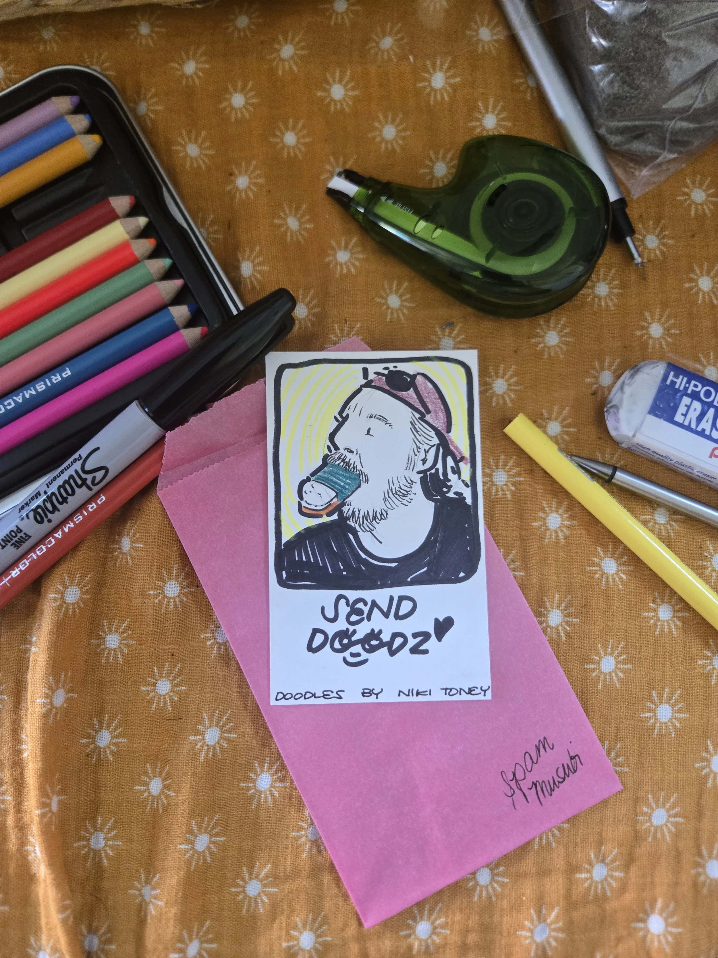 A collection of colorful markers, a small drawing of a man eating spam musubi, and a green tape dispenser on a polka-dotted orange tablecloth.