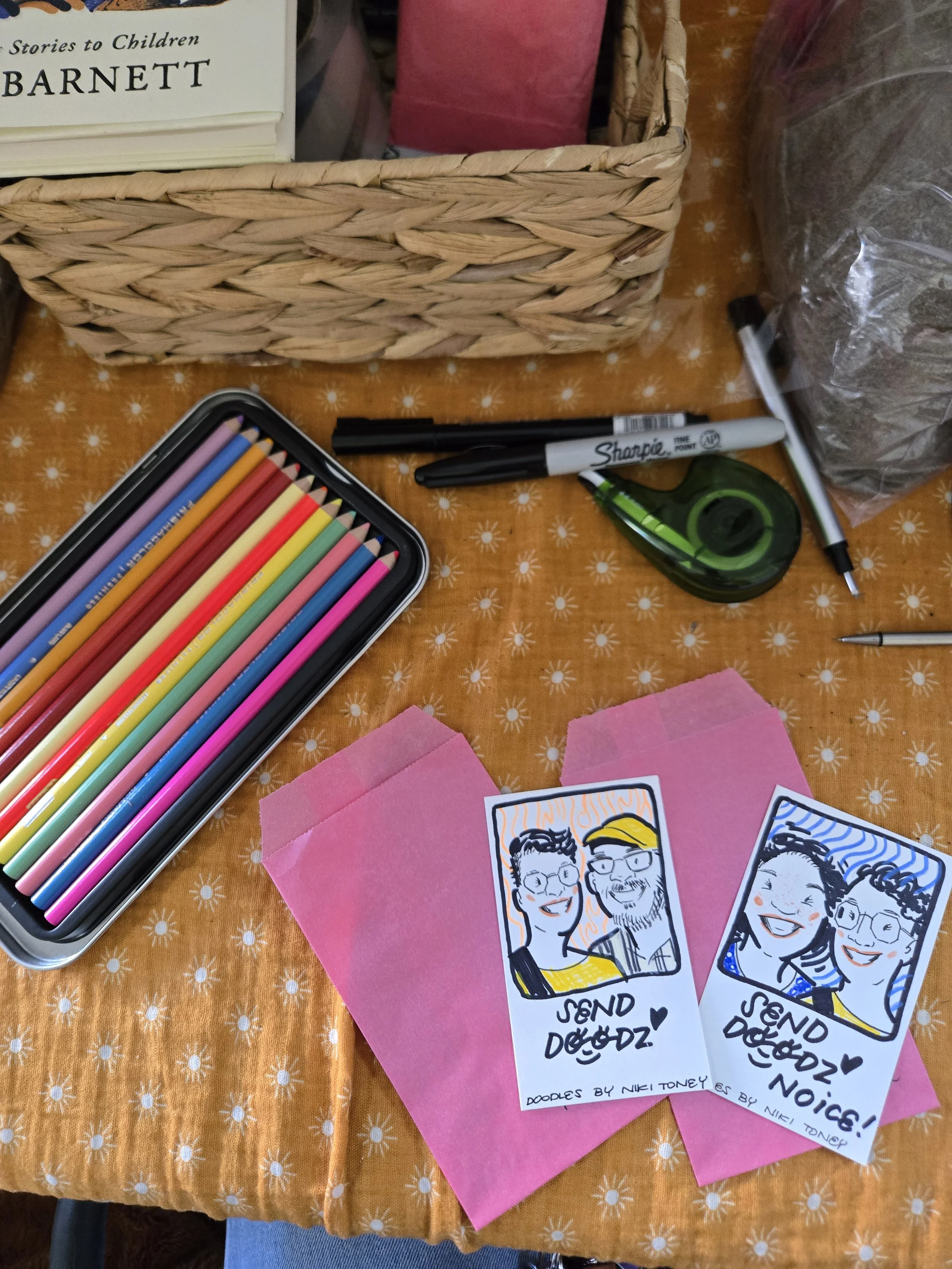 Colorful sketches of three people on small pink papers, with text that says 'Send Dödz!'. The sketches are in black and white with colorful details, and the paper has a hand-drawn style. There are markers, a tape measure, a black pen, and colored pencils on an orange table with a white flower pattern.