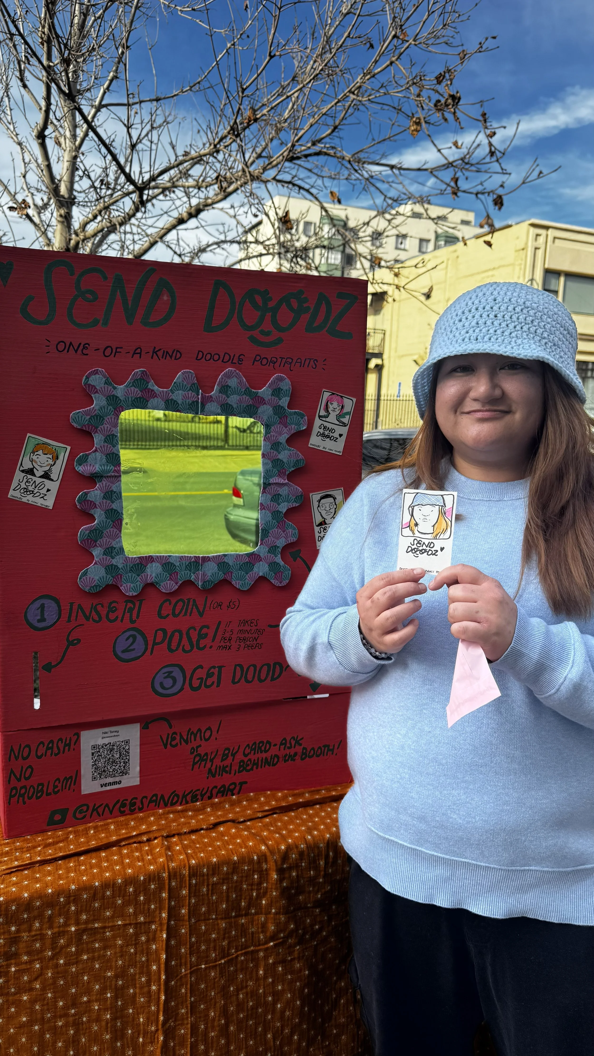 A woman holding a doodle portrait at an outdoor event, standing next to a pink booth titled "Send Doodz" with a sign providing instructions for the doodle portrait activity. The background features a leafless tree, a city building, and a blue sky.