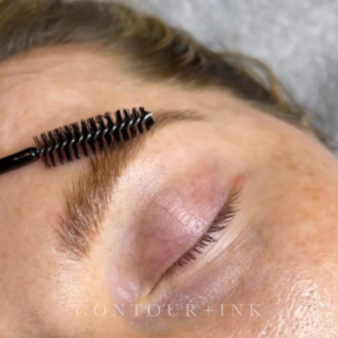 Close-up of a person's face with a black eyelash brush applying mascara to the upper eyelashes near the eyebrow.