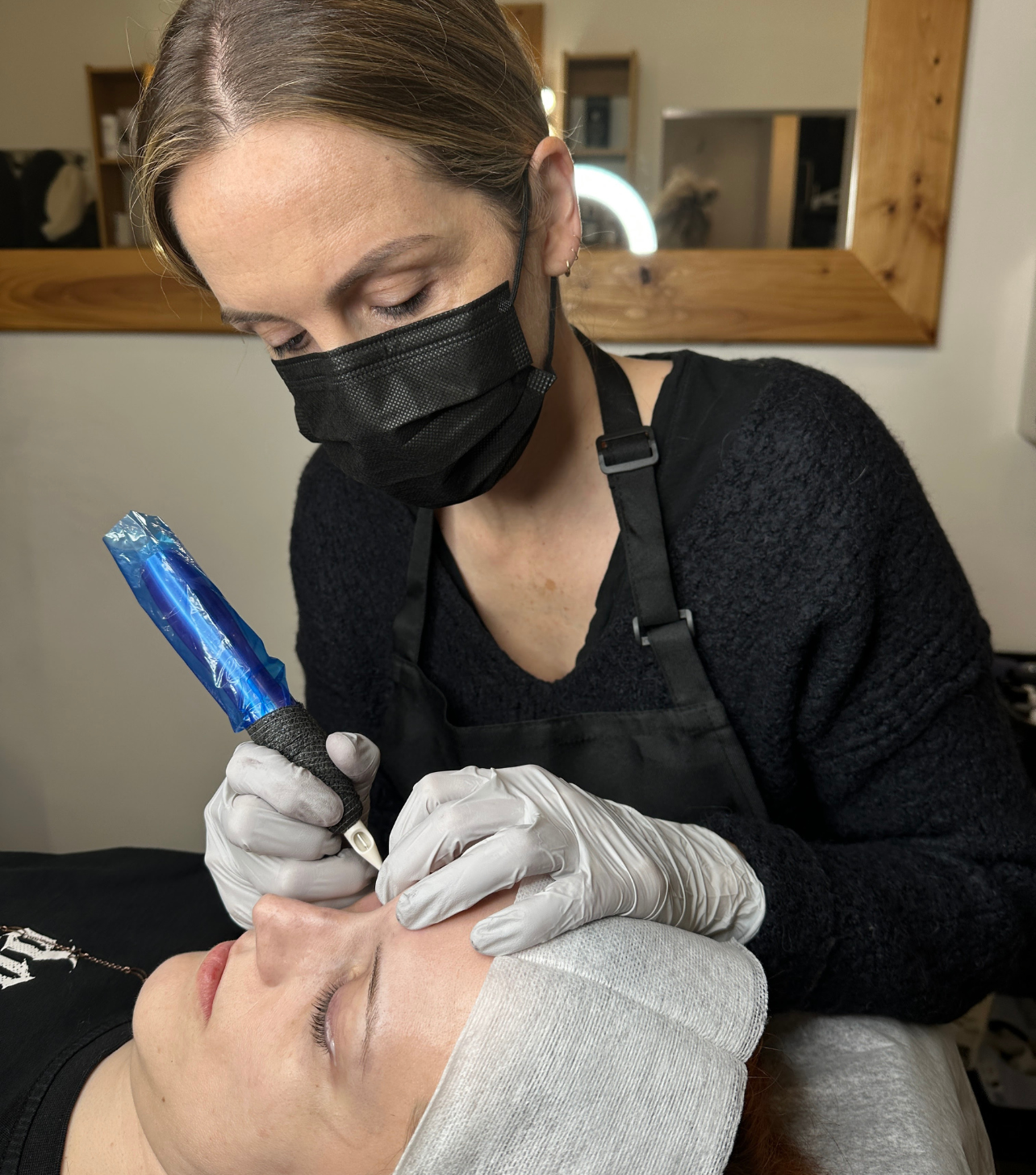 A woman receiving permanent makeup on her eyebrows from an artist wearing a black mask, gloves, and an apron in a salon.