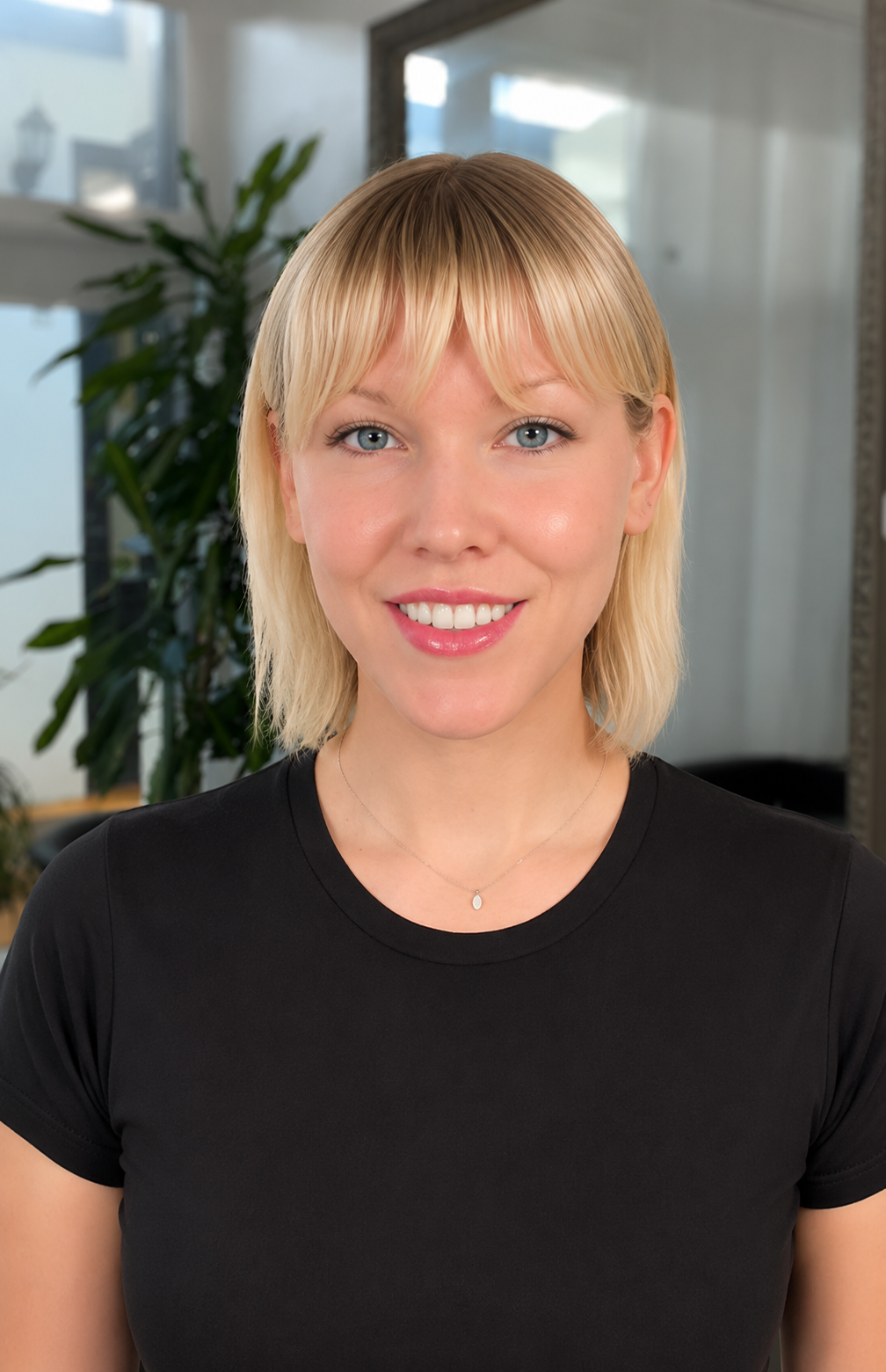 A young woman with blonde hair and blue eyes smiling at the camera, wearing a black t-shirt and a delicate necklace, in an indoor setting with a plant and window in the background.