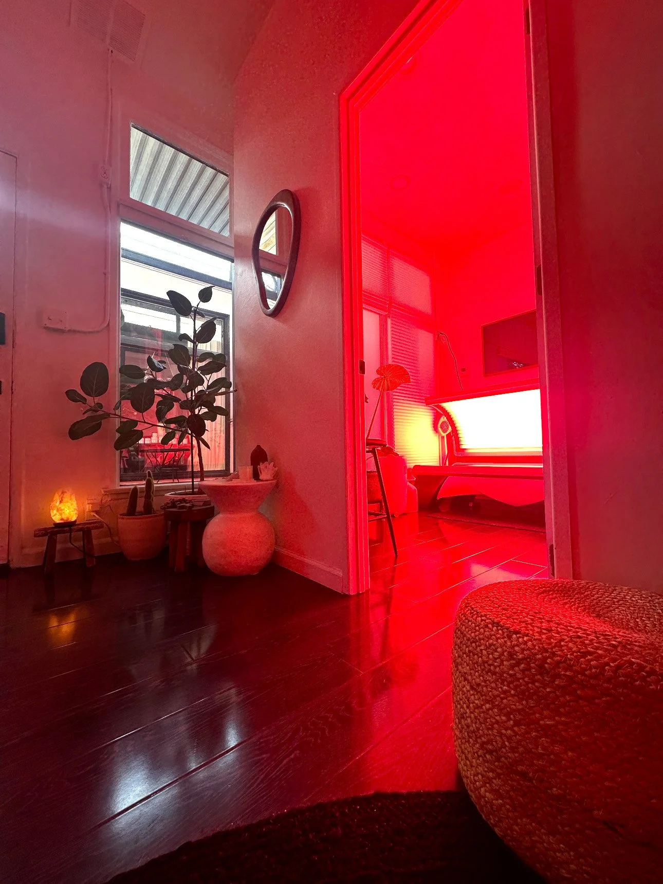 A room with red lighting, a doorway to another space, potted plants, a salt lamp, a mirror on the wall, and a window letting in natural light.