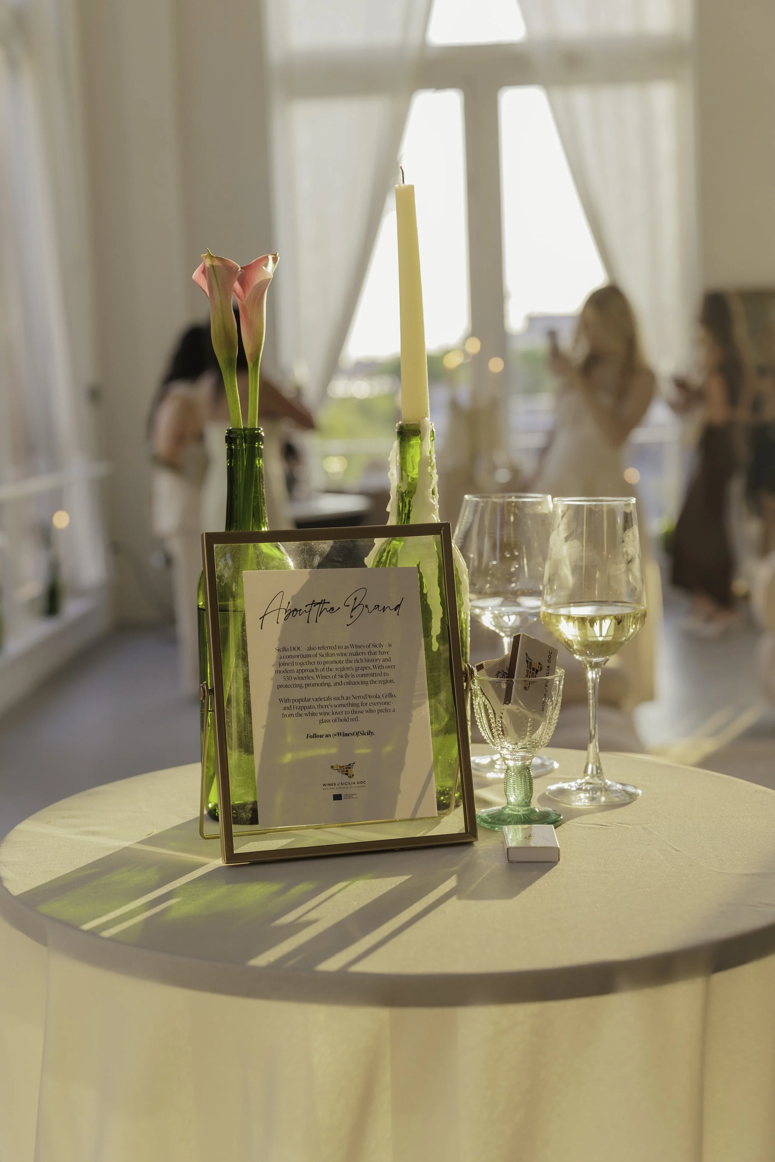 Elegant table setting with flowers, wine glasses, candles, and a framed card in a well-lit venue.