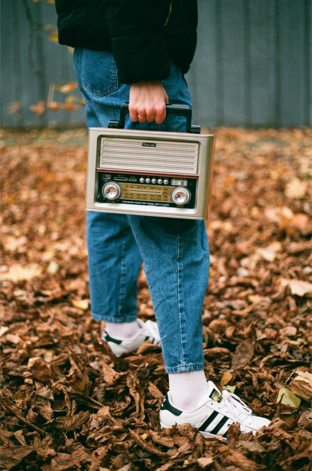 Person holding a vintage radio, standing on dry autumn leaves, wearing blue jeans and white sneakers with black stripes.