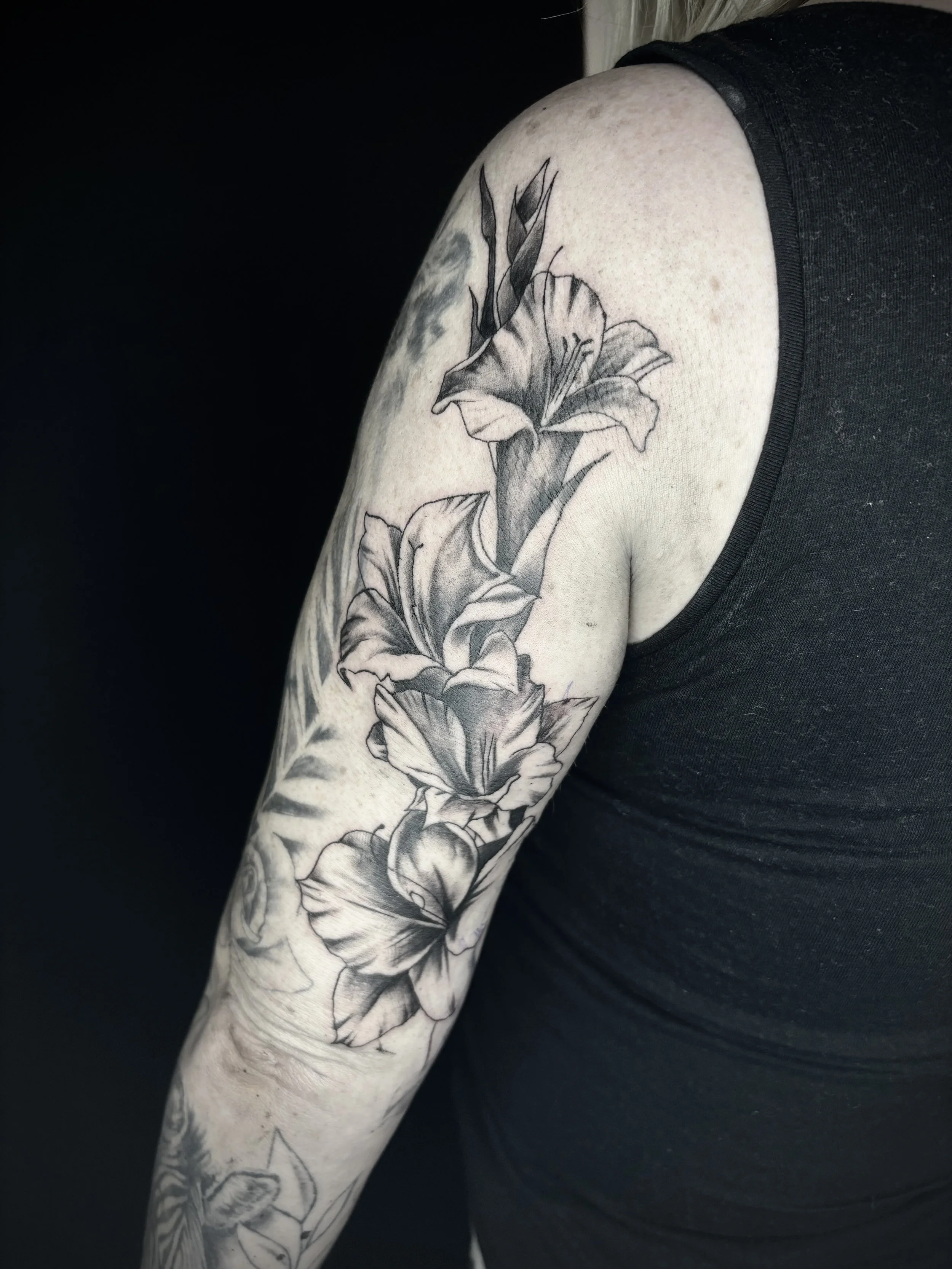 Black and white tattoo of several large flowers, likely lilies, on a person's upper arm and shoulder.