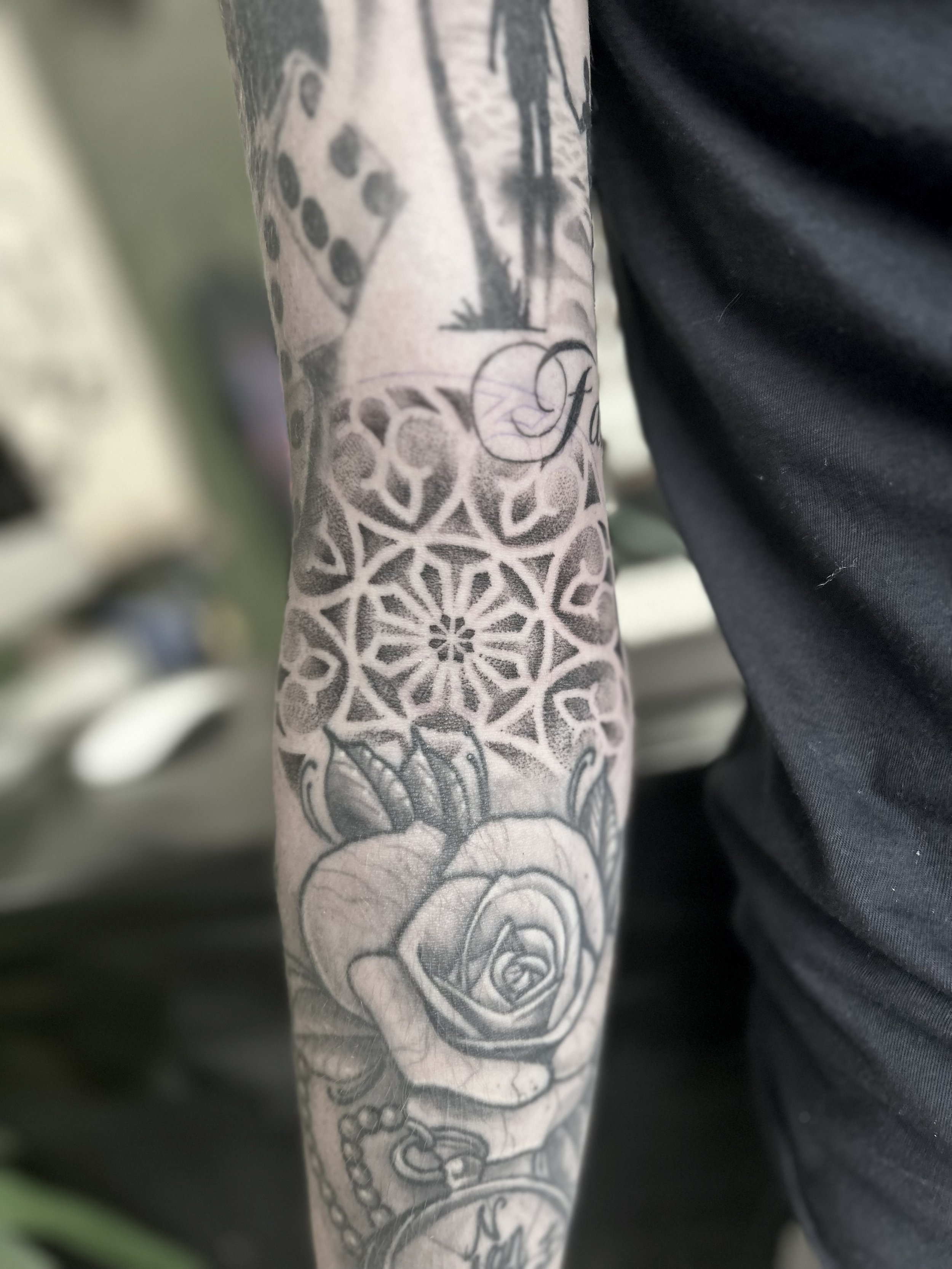 Close-up of a tattoo sleeve on a person's arm, featuring a detailed rose held by a hand, intricate geometric patterns, and other decorative elements.