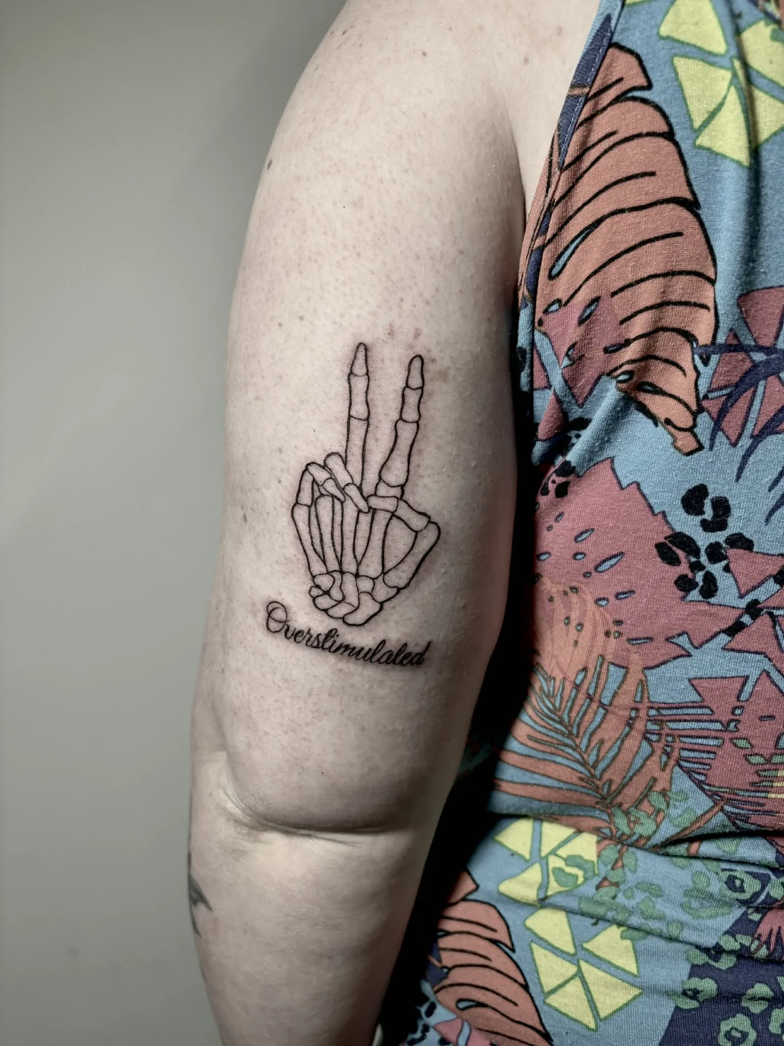 Tattoo of a skeleton hand making a peace sign, with the word 'Overstimulated' written below, on a person's upper arm.