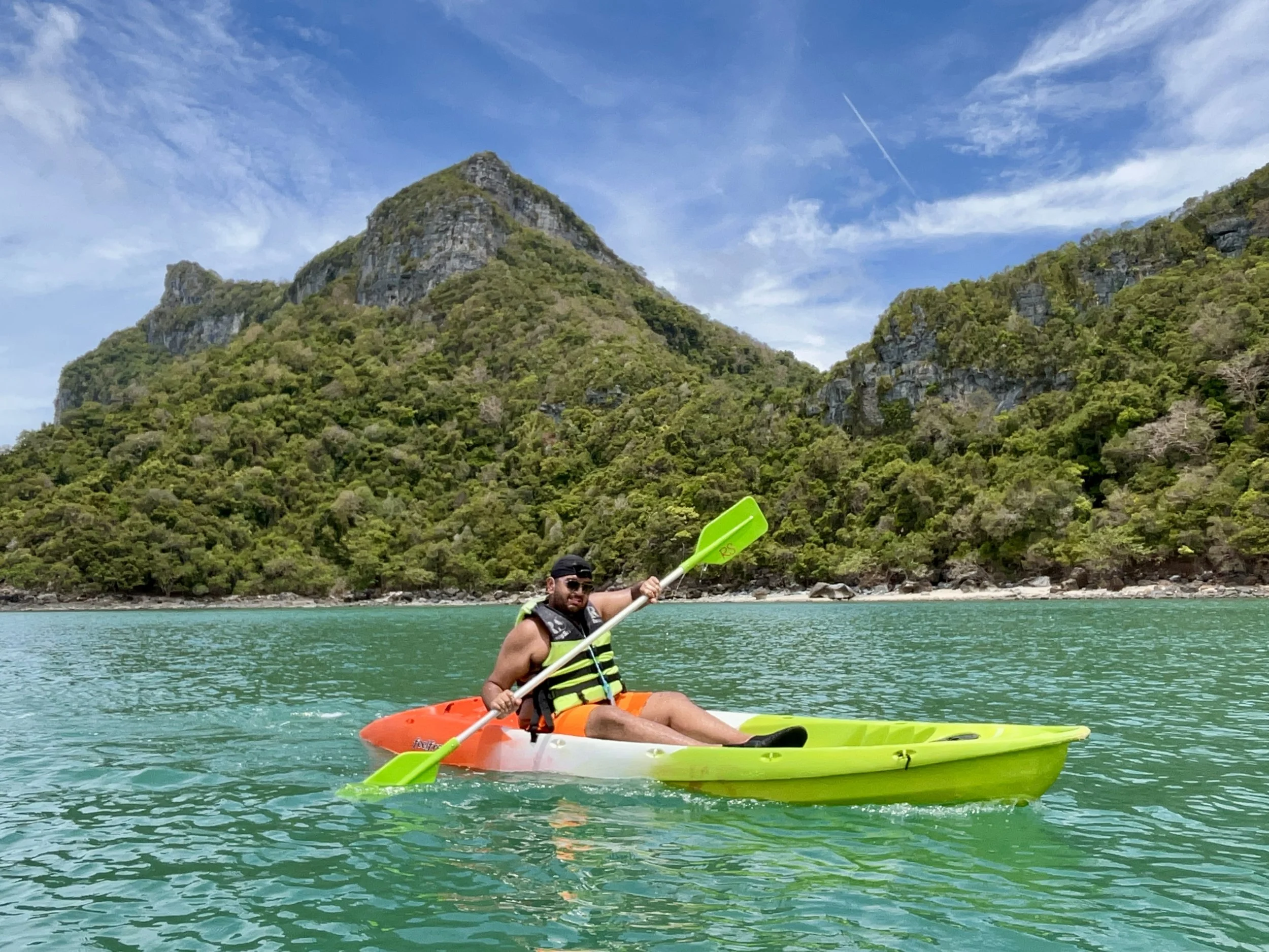 A man in sunglasses and a life jacket kayaking in a turquoise lake with lush green mountains and a partly cloudy sky in the background.