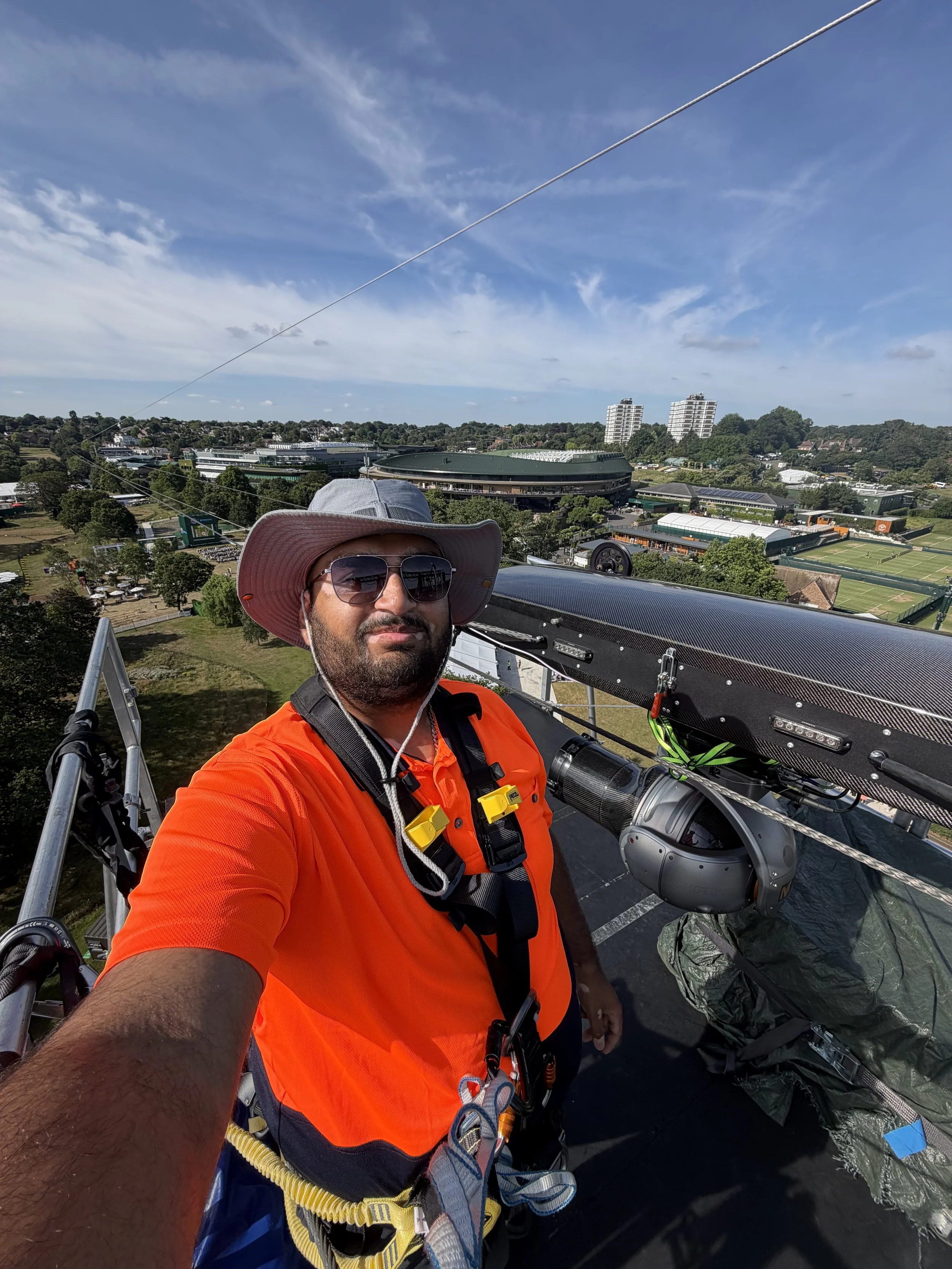 A man wearing a large hat, sunglasses, an orange safety vest, and harness, standing on a metal platform high above the ground, with a cityscape including Wimbledon tennis courts and tall buildings in the background.
