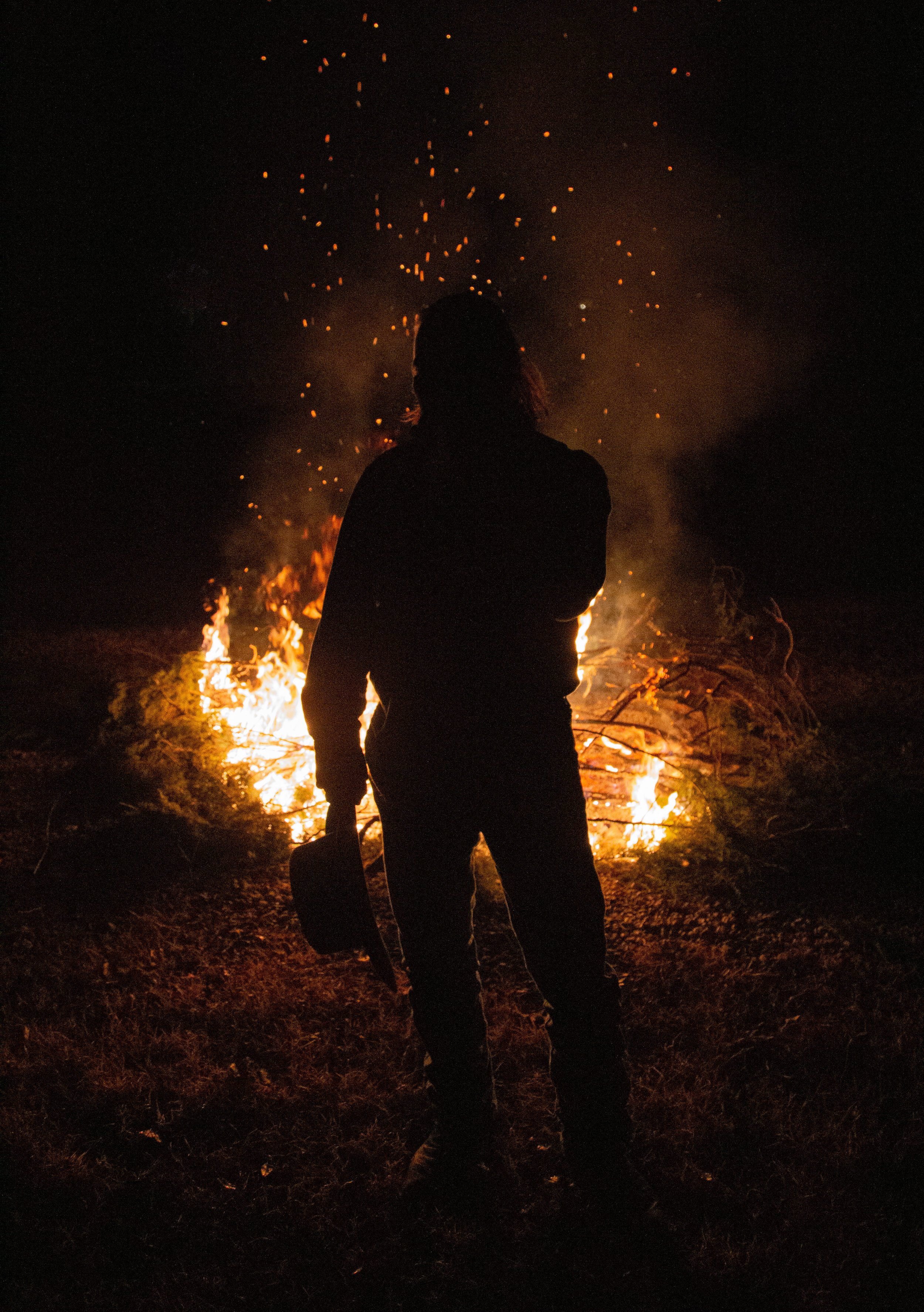 Silhouette of a person holding a hat or helmet, standing in front of a large bonfire at night with orange flames and sparks.