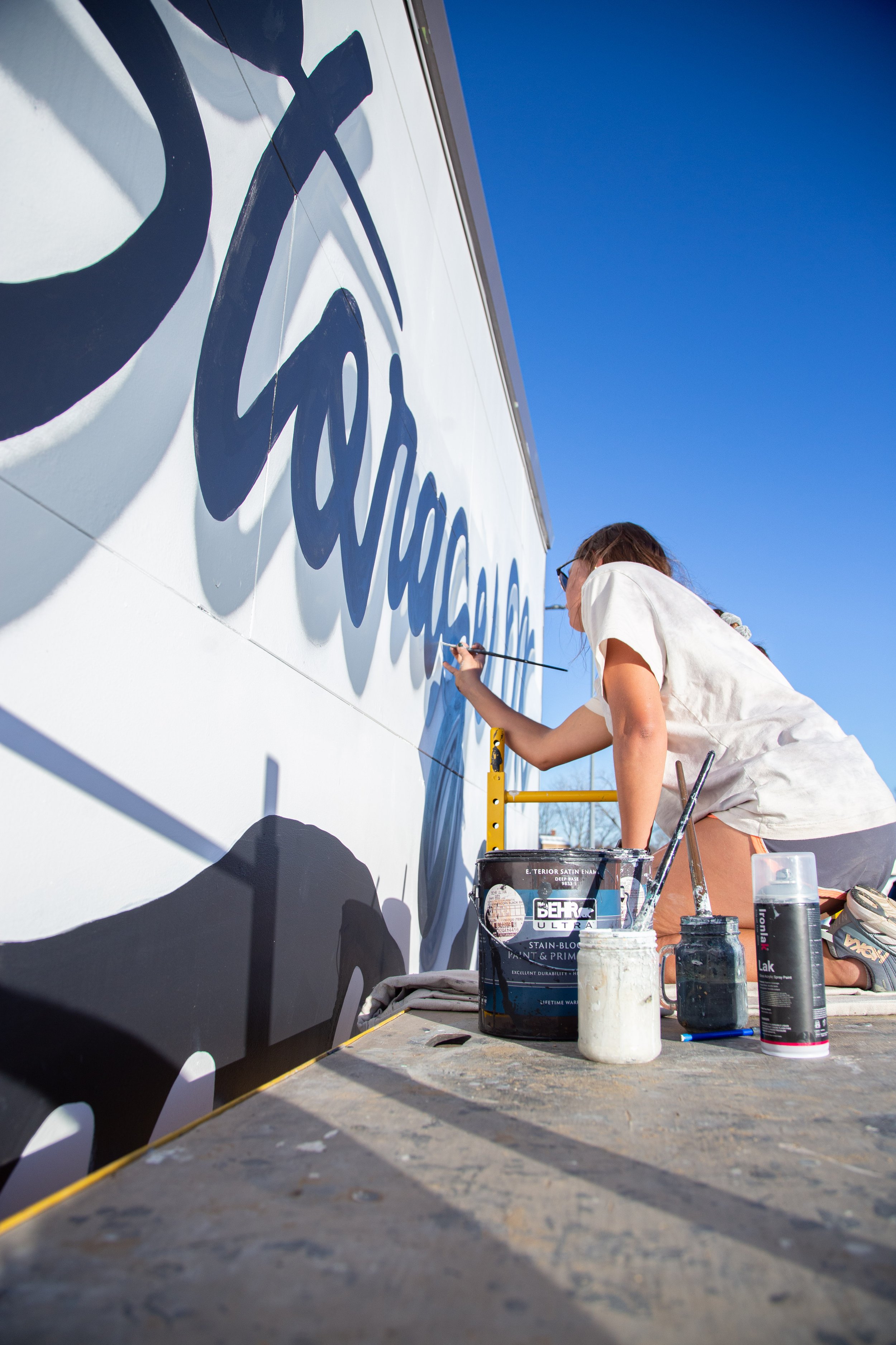 A woman painting a logo on a white exterior wall under a clear blue sky, with paint supplies and brushes nearby.