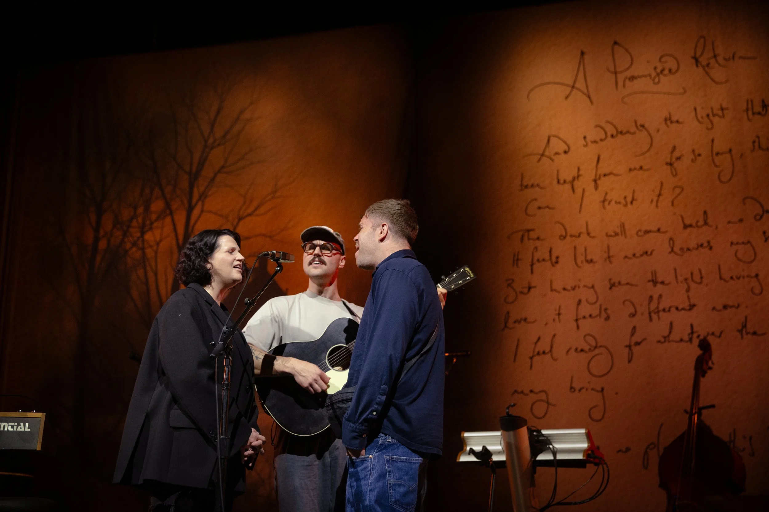 Three musicians performing on stage, one of them the artist, Haffway, with a dark background and tree silhouette, and handwritten text projected on the wall. They are on stage at The Ryman in Nashville. 