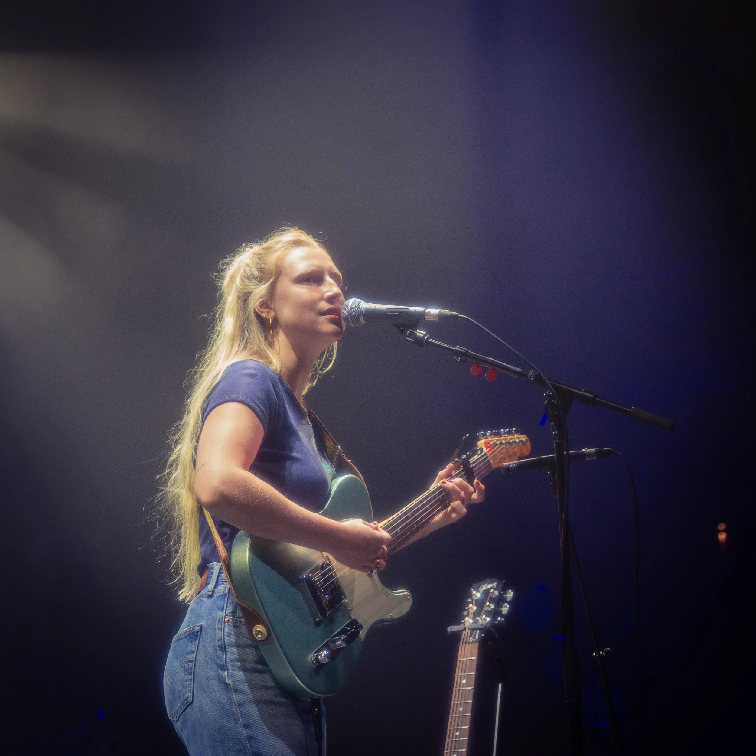 The musician Billie Marten, playing an electric guitar and singing into a microphone on stage, with dark background and stage lighting.