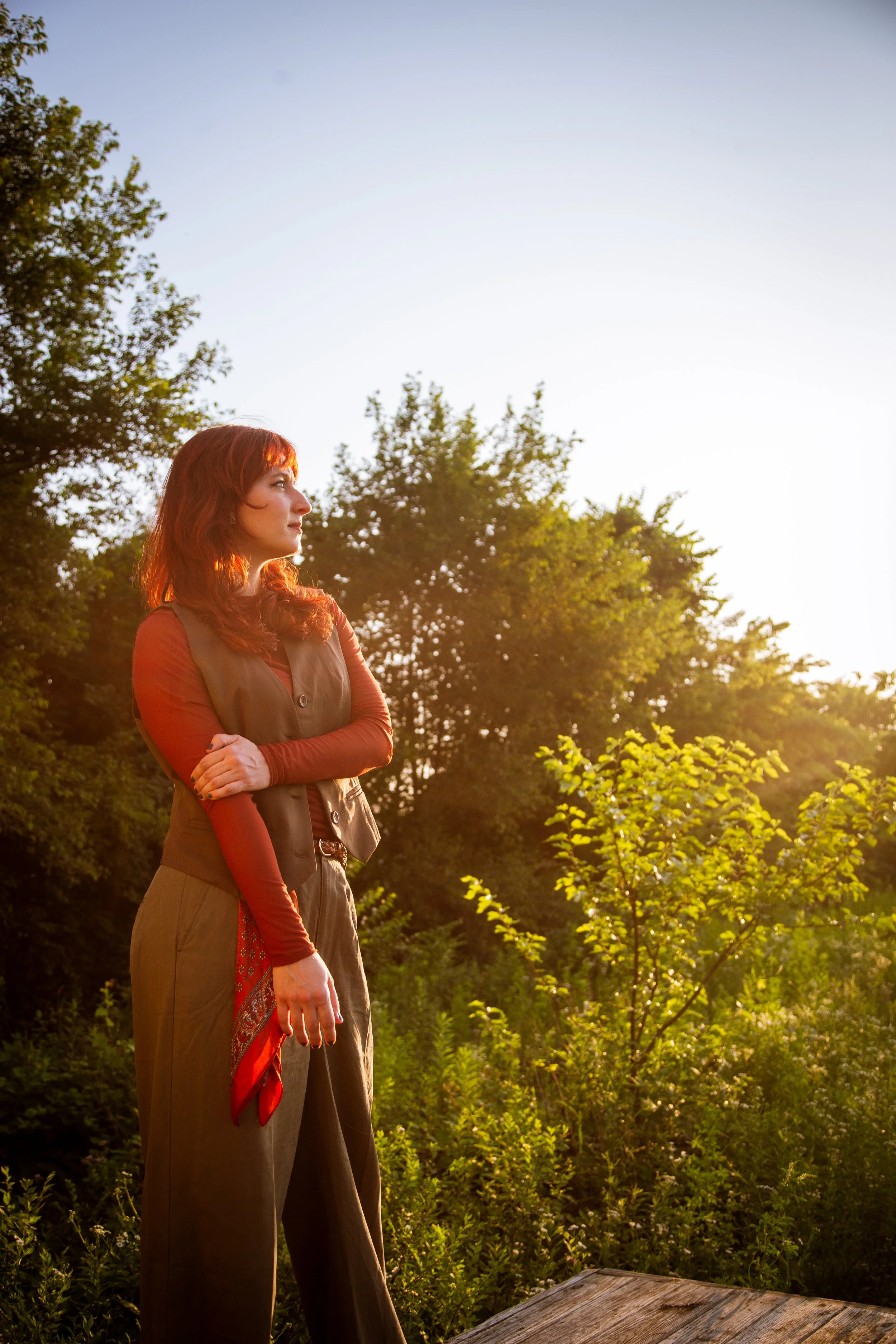 A woman with red hair standing outdoors at sunset, surrounded by green trees and foliage, looking to the side with crossed arms.