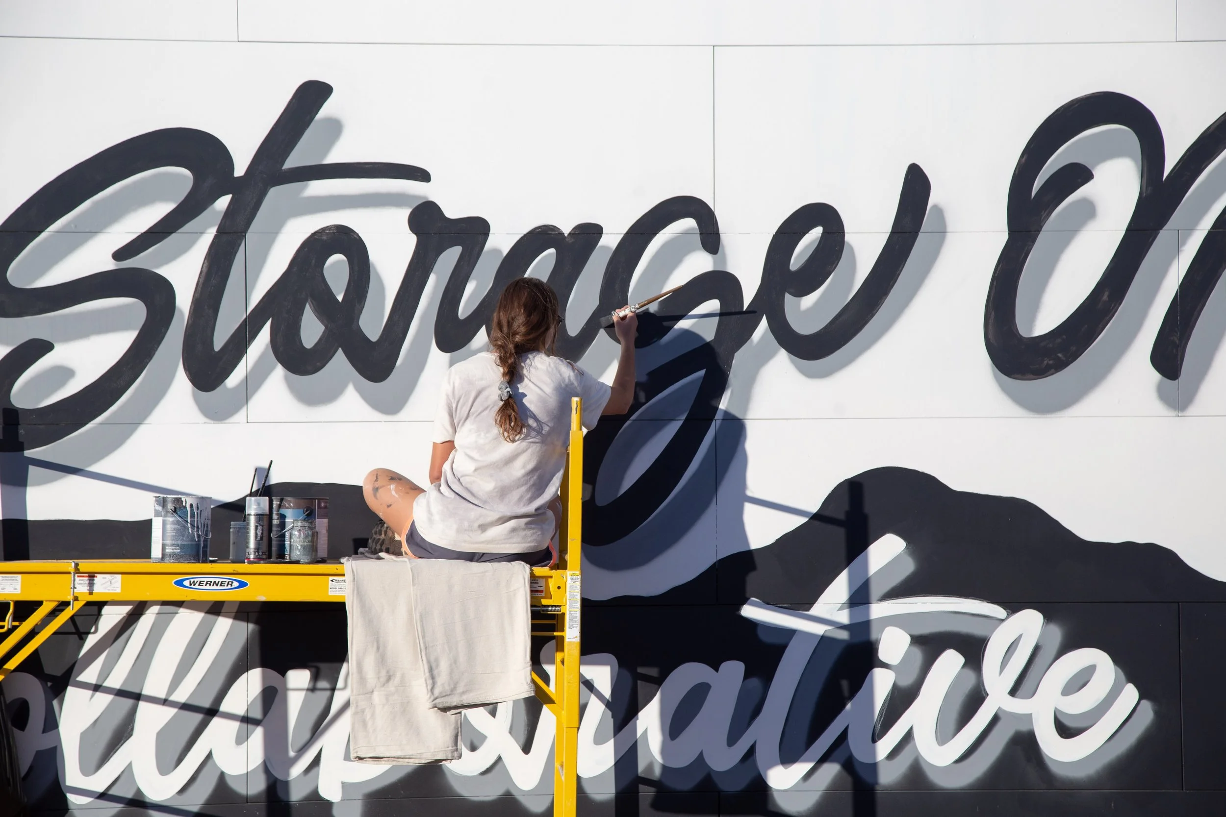 A woman painting a mural on a wall reads 'Show me the way to celebrate'. She is sitting on a yellow lift with cans of paint nearby.