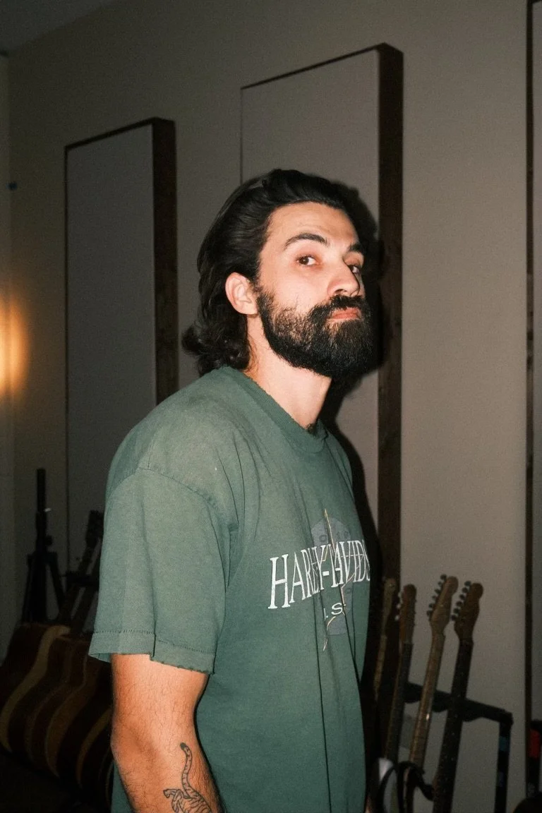 A man with long dark hair, a beard, and a mustache, wearing a green T-shirt with a Harley-Davidson logo, standing in a room with guitars in the background.