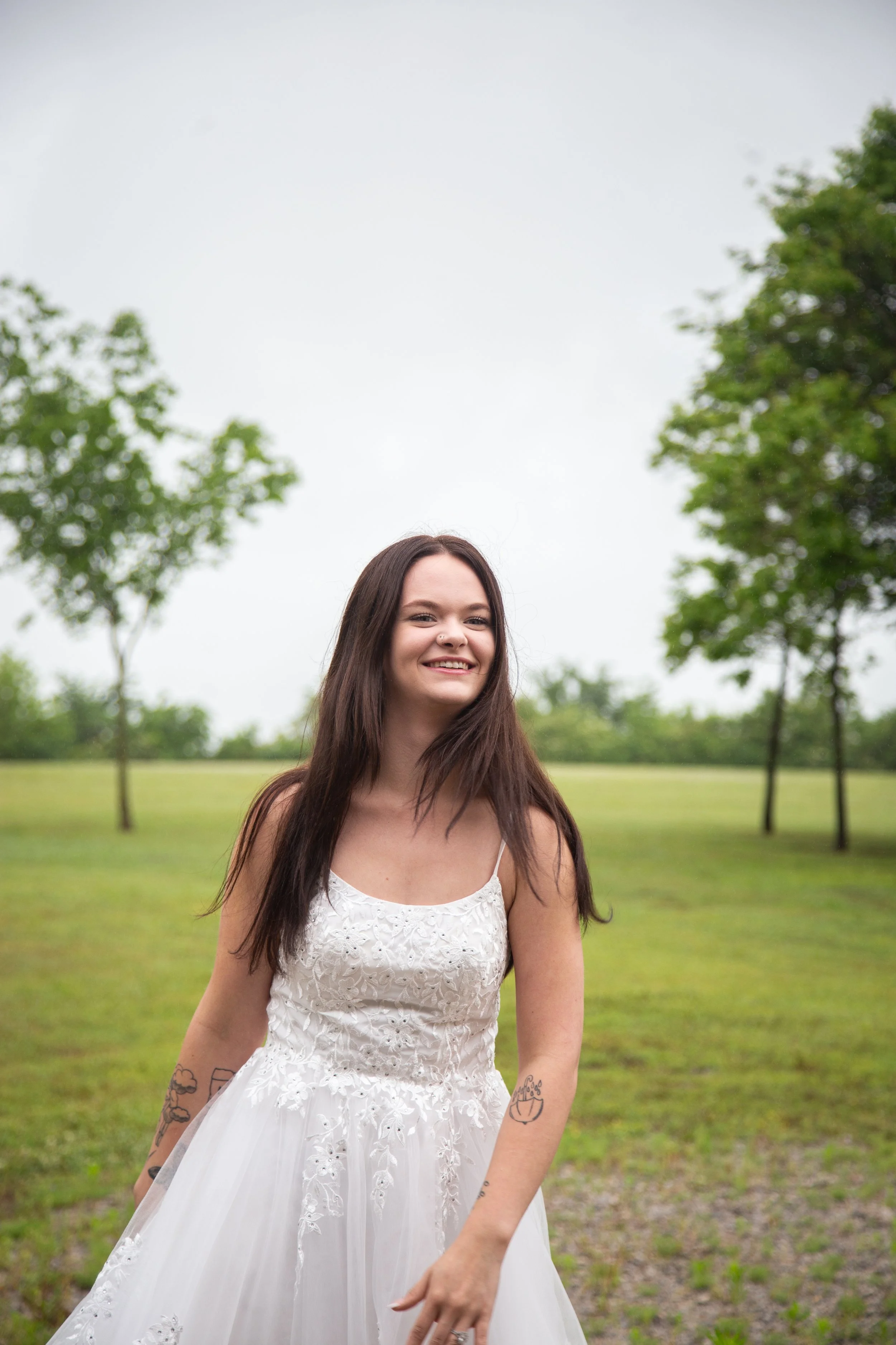 A woman in a white dress standing outdoors on a green grassy field with trees in the background, smiling.