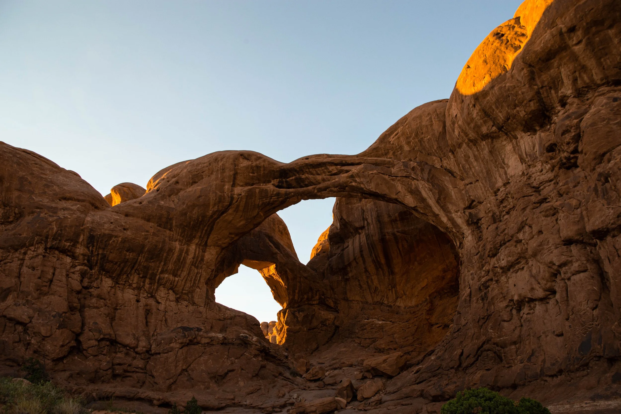 Natural sandstone rock formation with arch and a small heart-shaped opening, illuminated by sunlight at sunset or sunrise, in a desert landscape with sparse greenery.