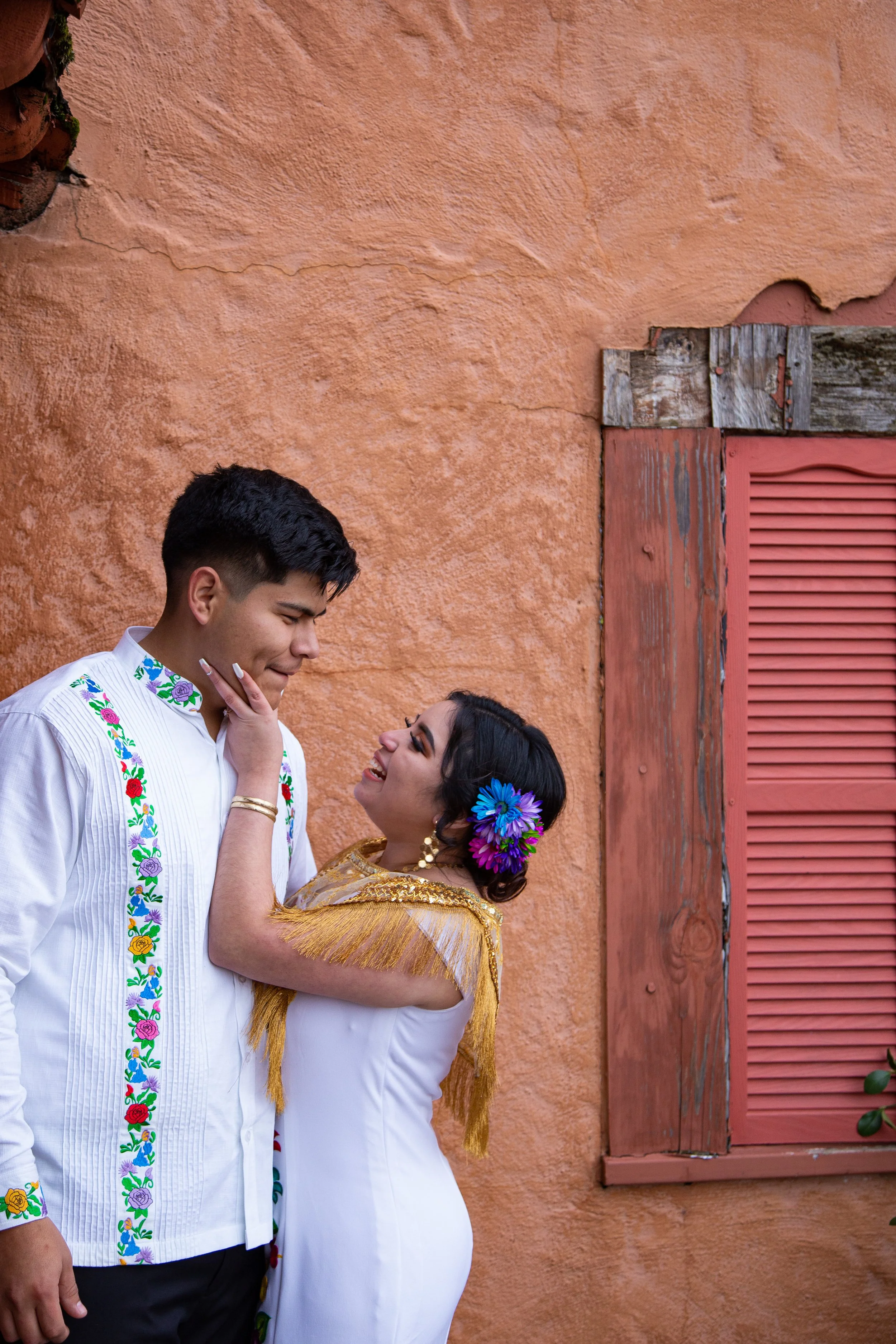 A couple dressed in traditional Mexican clothing standing close to each other, smiling, with a textured orange wall and pink window shutter in the background.