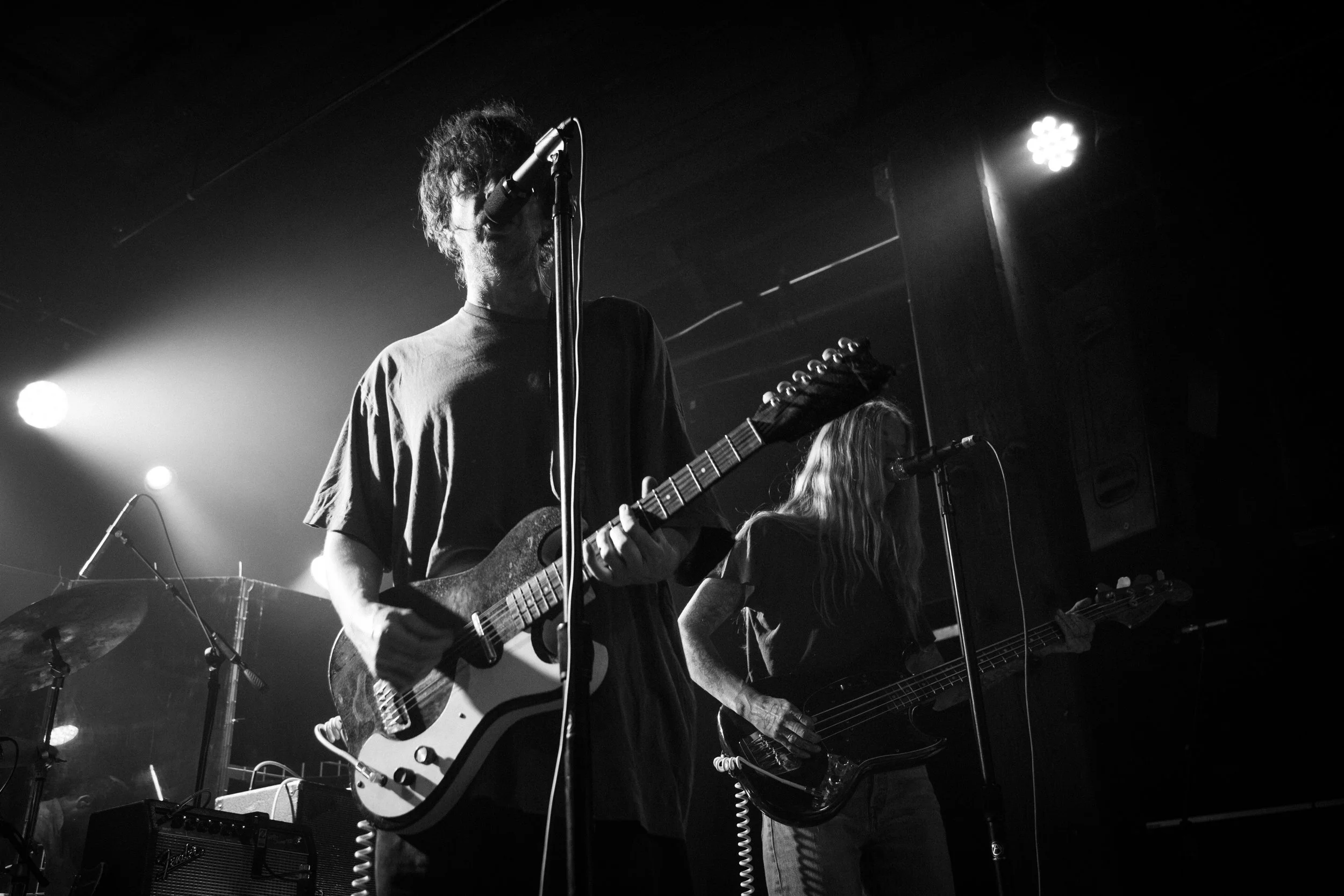 Black and white photo of the Oklahoma based band, Broncho, on stage playing an electric guitar, with a microphone in front of him, and another musician partially visible beside him, with stage lights behind and equipment on stage.