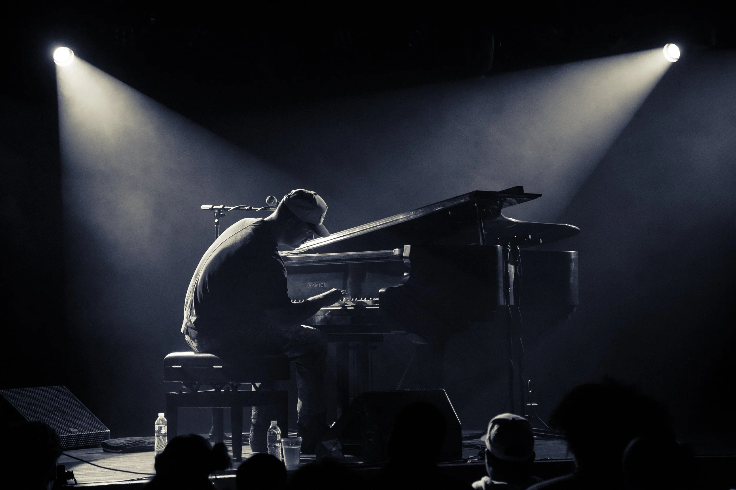 A person wearing a cap and glasses playing a grand piano on a dimly lit stage with spotlights overhead.