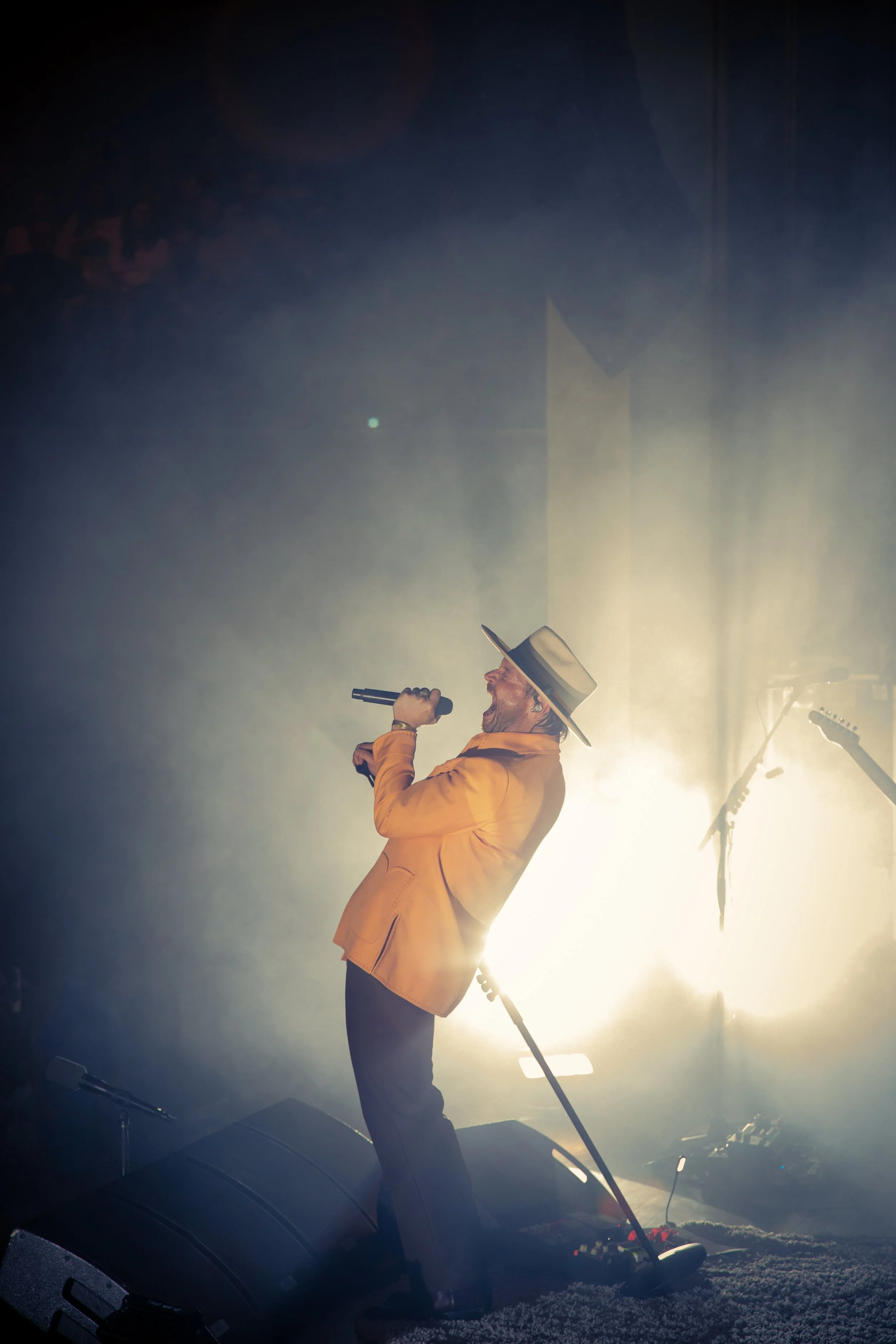 A man wearing a yellow jacket and a wide-brimmed hat singing into a microphone on stage with bright lights behind him.