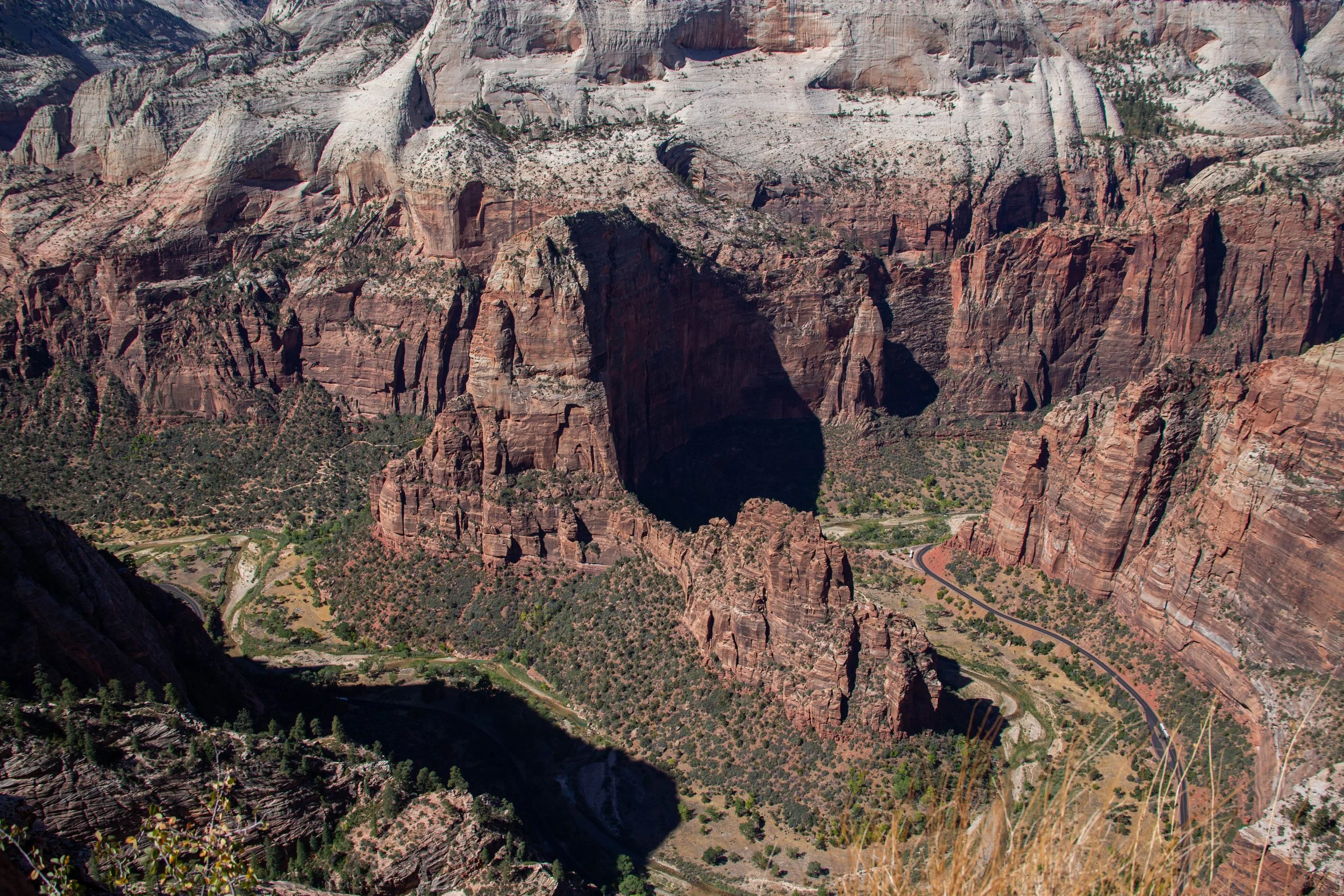 A view of the Grand Canyon showing steep, layered red rock cliffs, a deep shadowed canyon, and a winding road at the bottom with some sparse green vegetation.