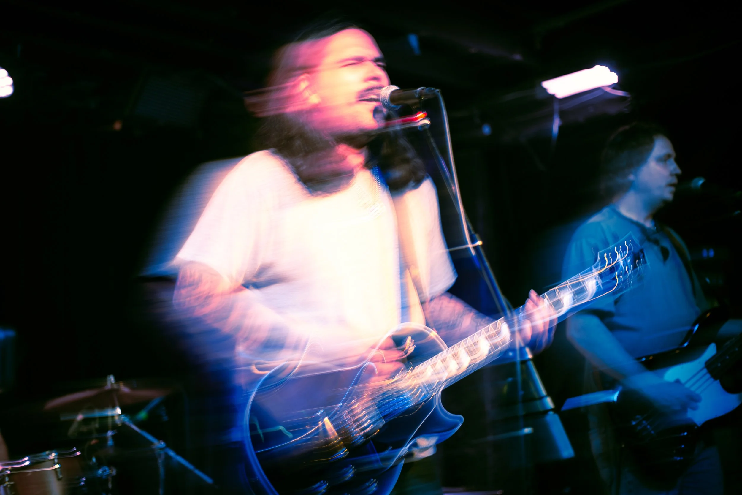 Blurred image of musician, Glom, singing into a microphone and playing a guitar in a dark setting with stage lights.