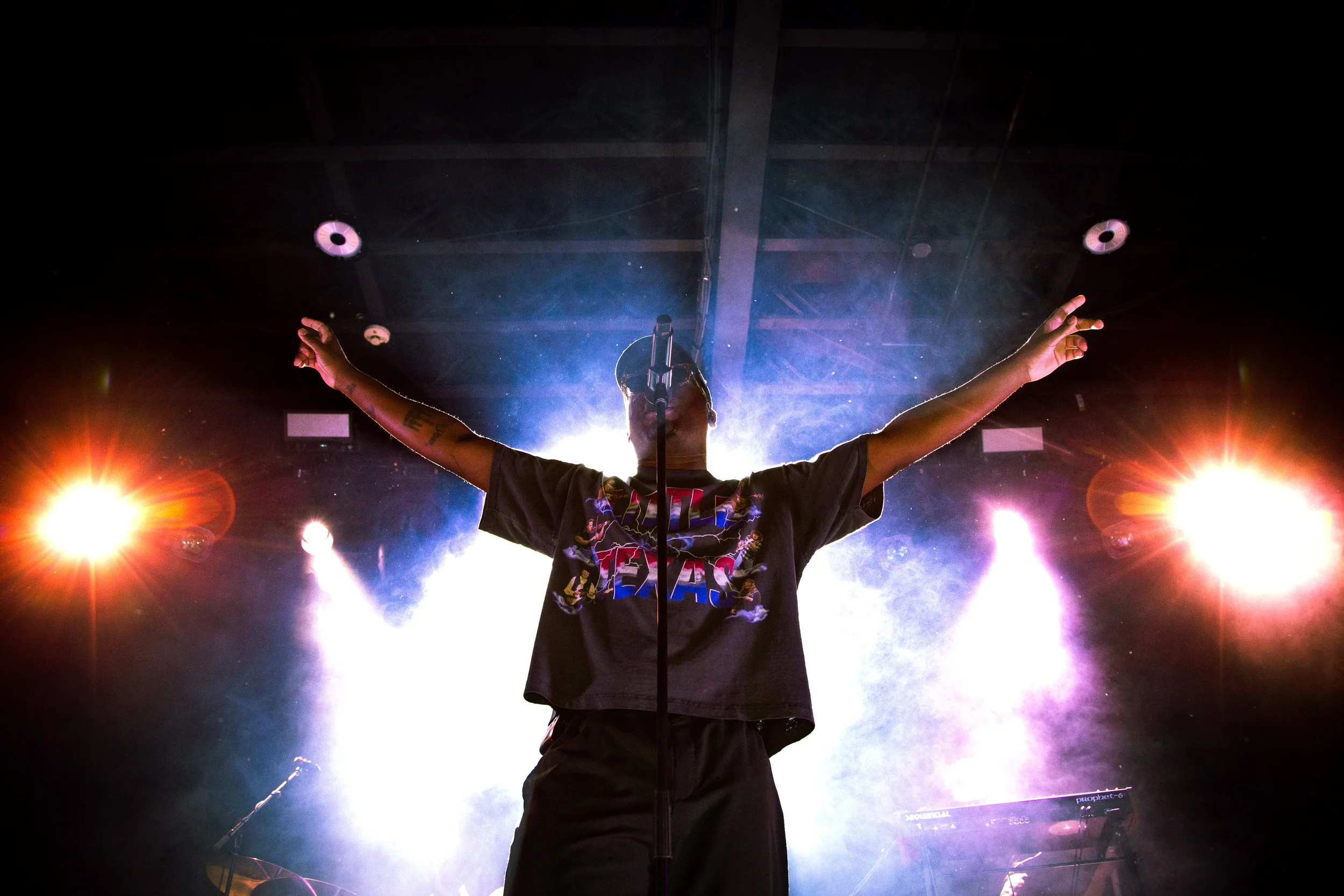 Performer on stage with arms outstretched in front of bright stage lights and smoke.