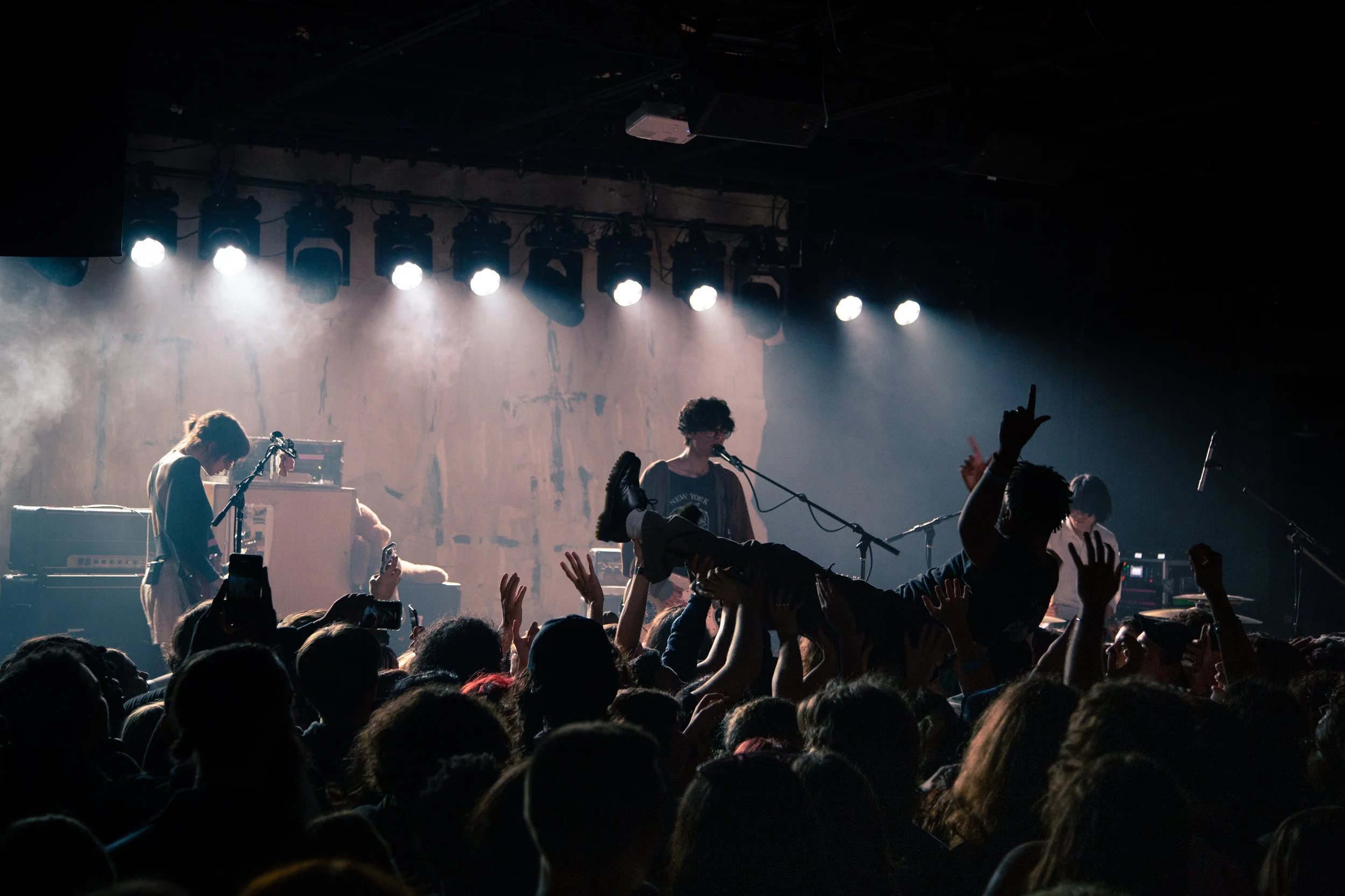Music concert with band playing on stage, audience crowd in front, some hands raised and crowd surfing, stage lighting overhead.