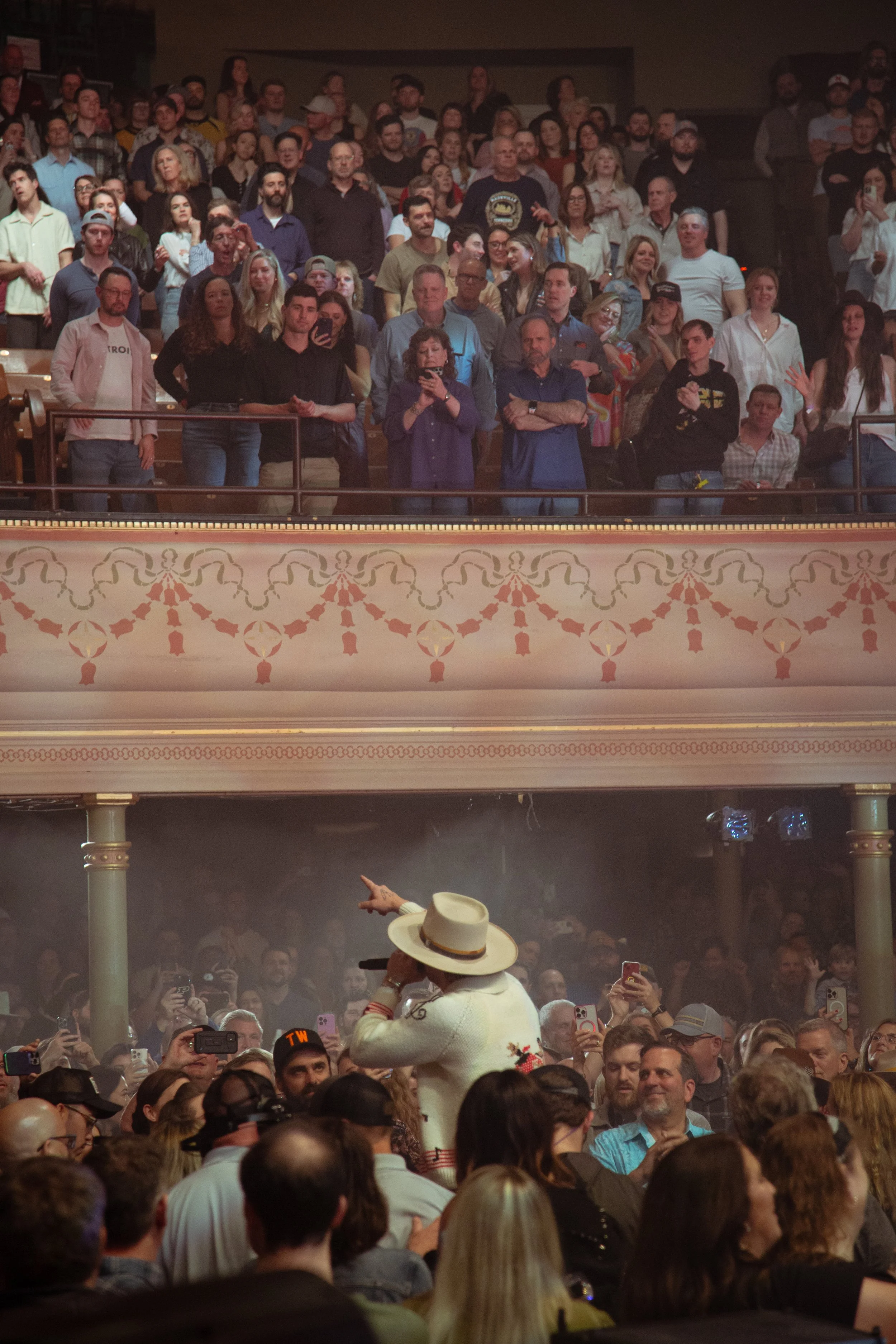A performer wearing a white hat and white outfit sings into a microphone on stage, with a large audience in the auditorium watching.