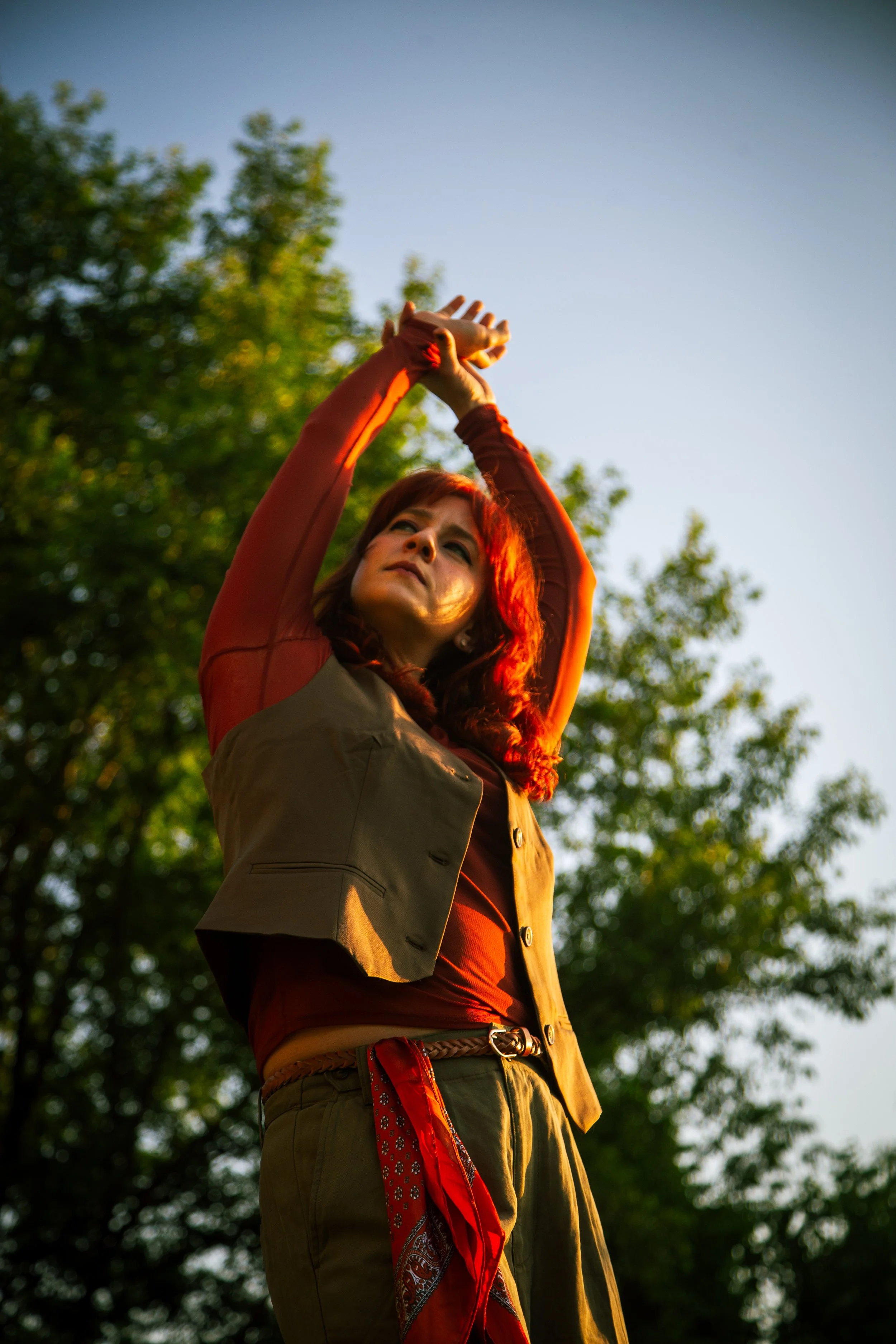 A woman with red hair stretching her arms upward outdoors during sunset, with green trees in the background.