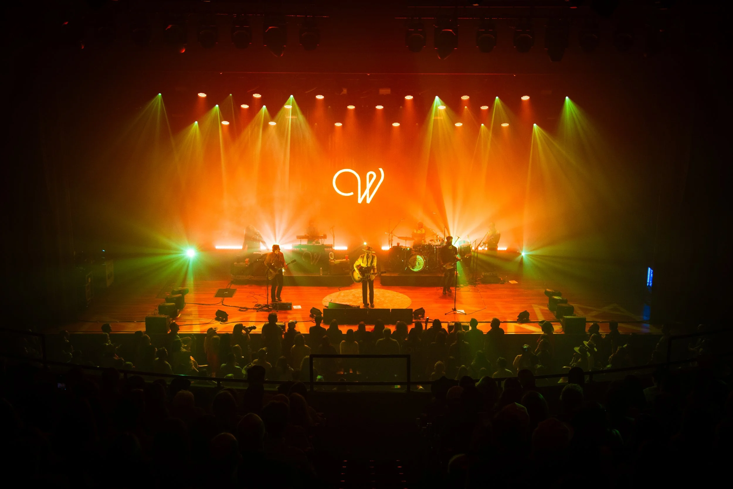 A band performing on stage with colorful lights and a large illuminated letter 'W' in the background, and an audience watching in front.