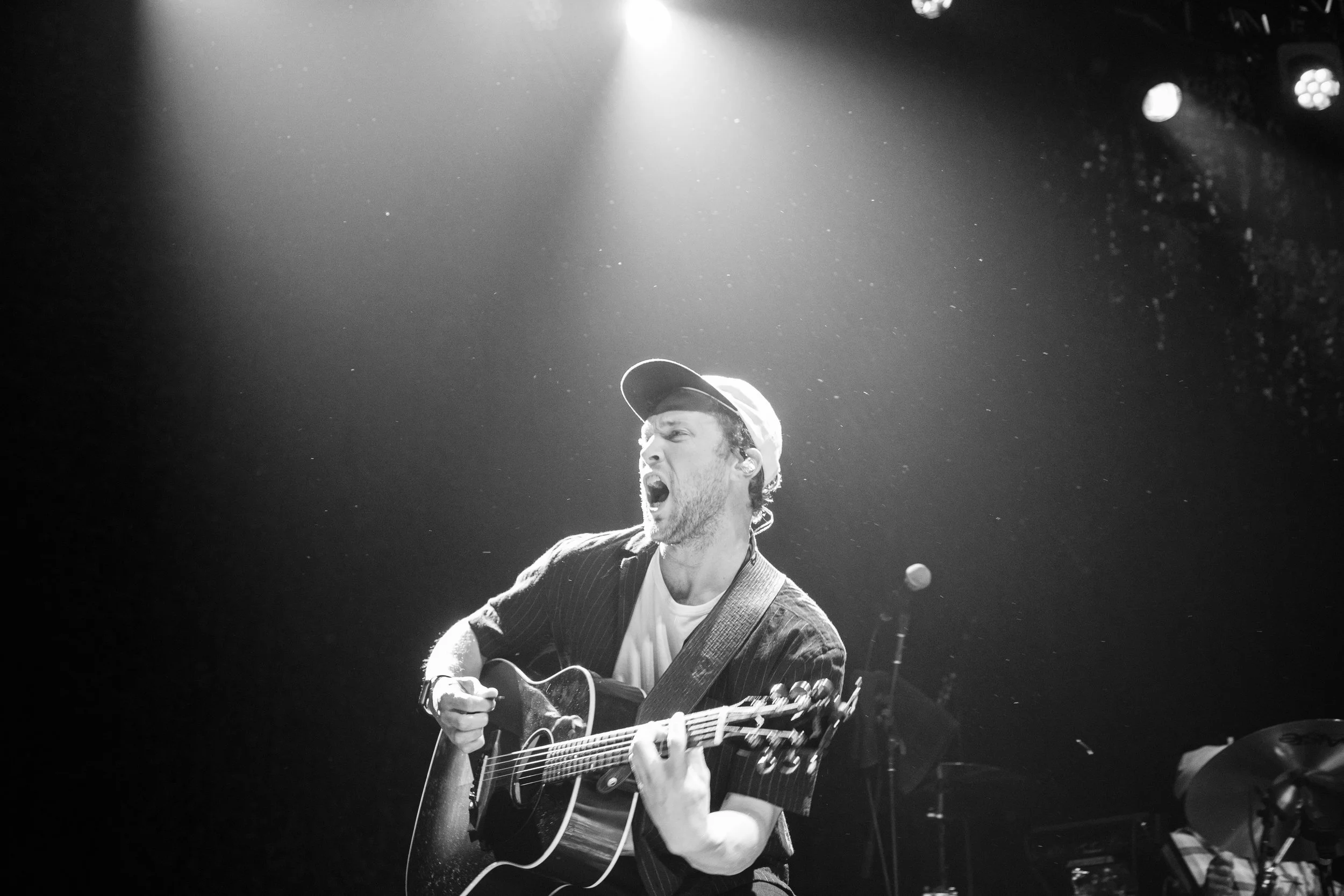 A man singing passionately and playing an acoustic guitar on stage, illuminated by bright stage lights in black and white.