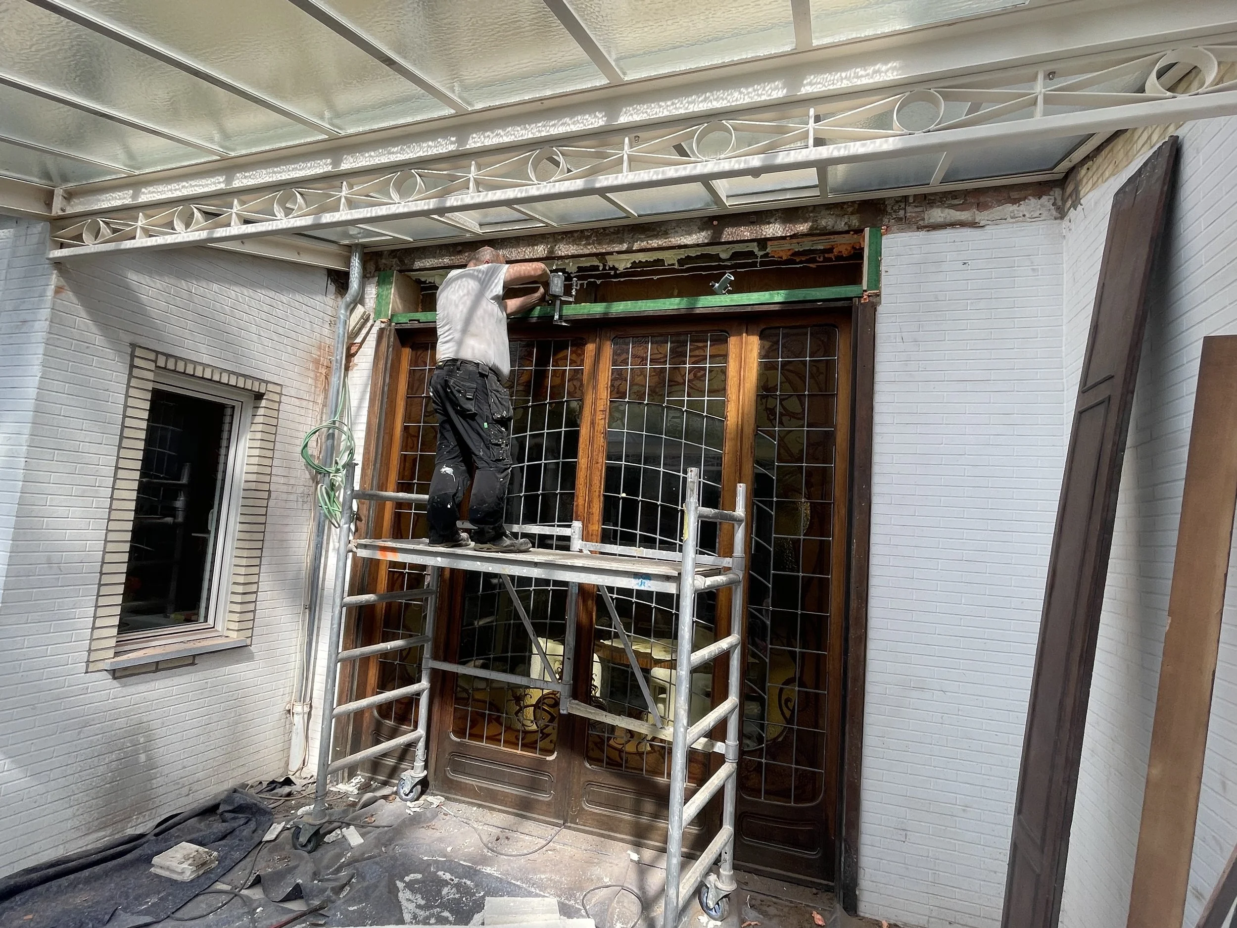 Een man werkt aan een houten voordeur met een metalen rooster, hij staat op een steiger. Bouwmaterialen en gereedschap liggen op de vloer voor de deur.