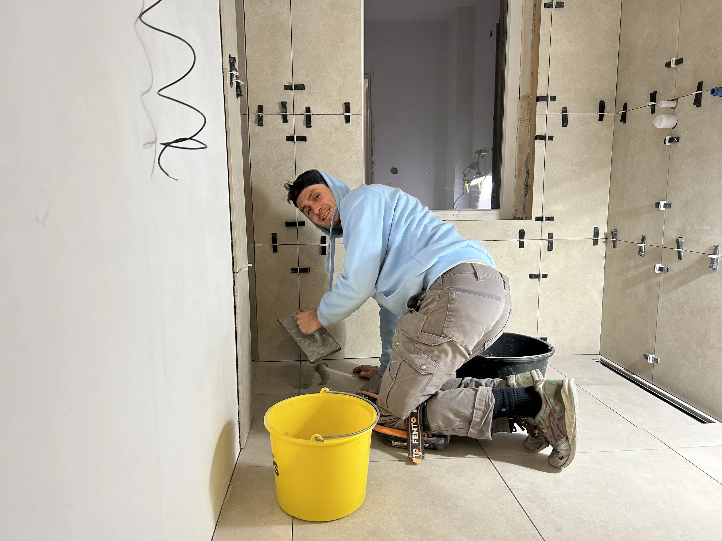 Man aan het tegelzetten in een ruimte, hij knielt op de vloer met een troffel in de hand, omringd door bouwmaterialen.