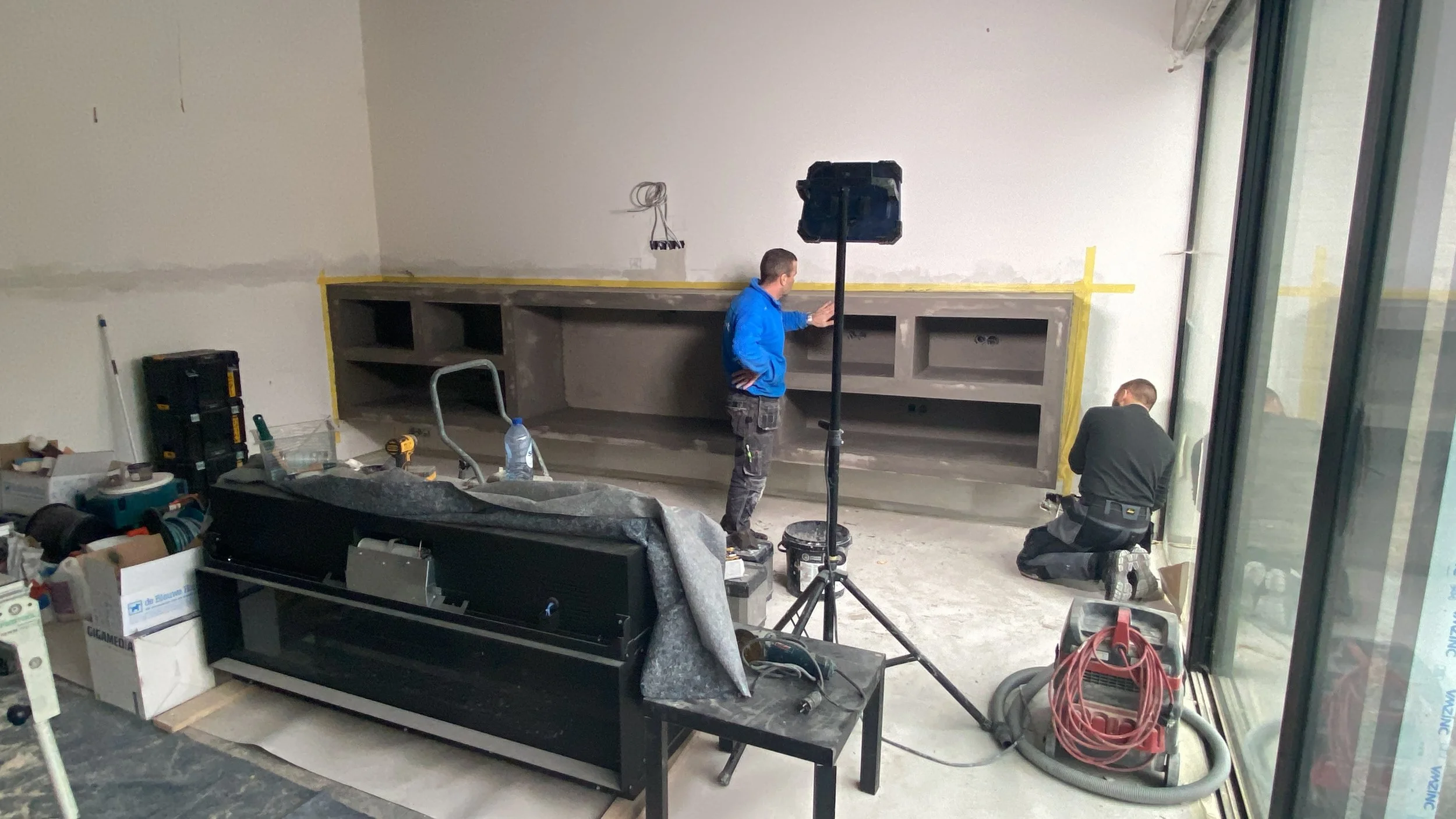 Binnenkant van een kamer onder constructie, met twee mannen die werken, bouwmaterialen, gereedschap en een grote zwarte machine.