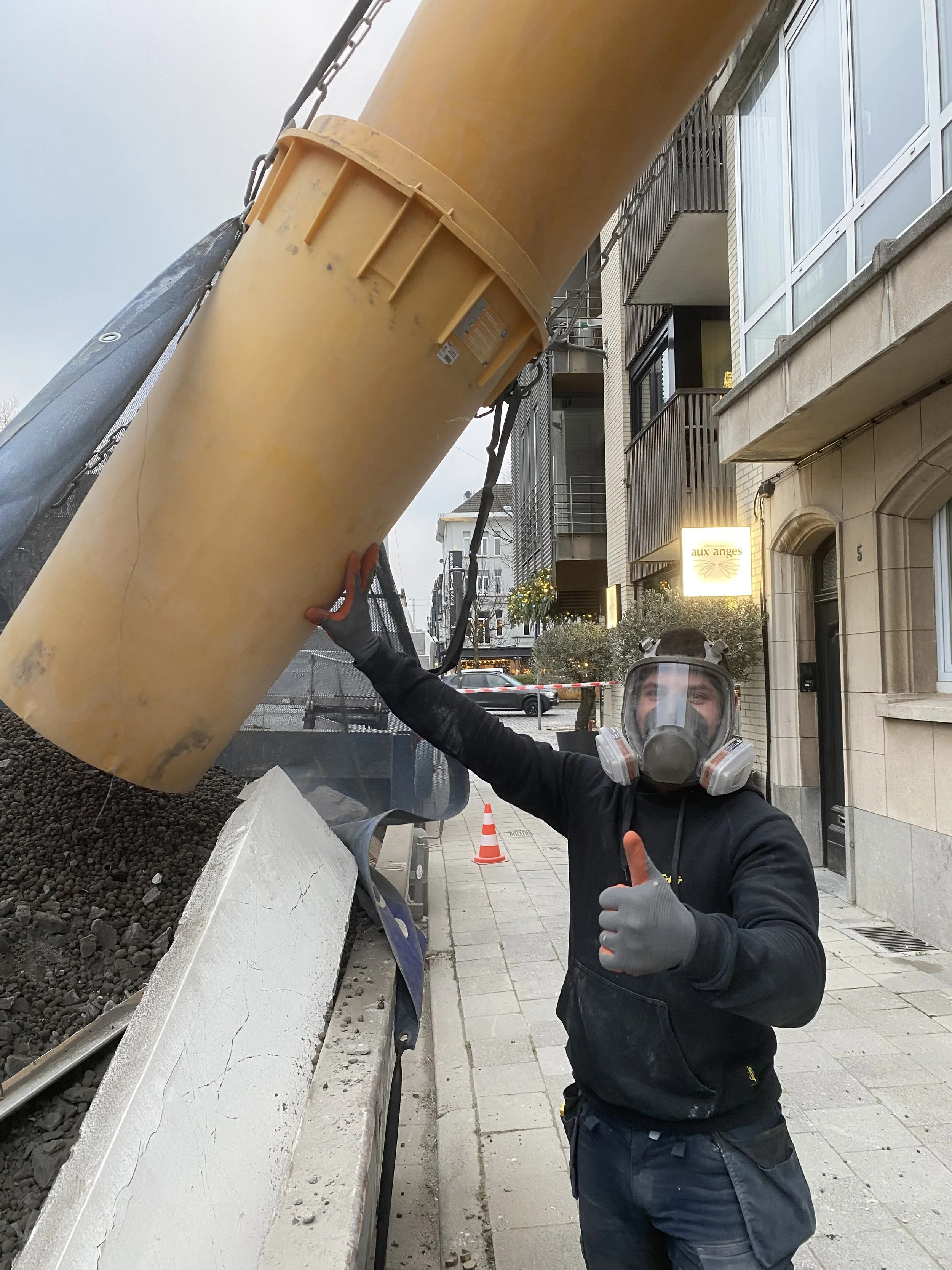 Een arbeider met beschermingsuitrusting geeft een duim omhoog terwijl hij een grote gele machine op de straat bedient.