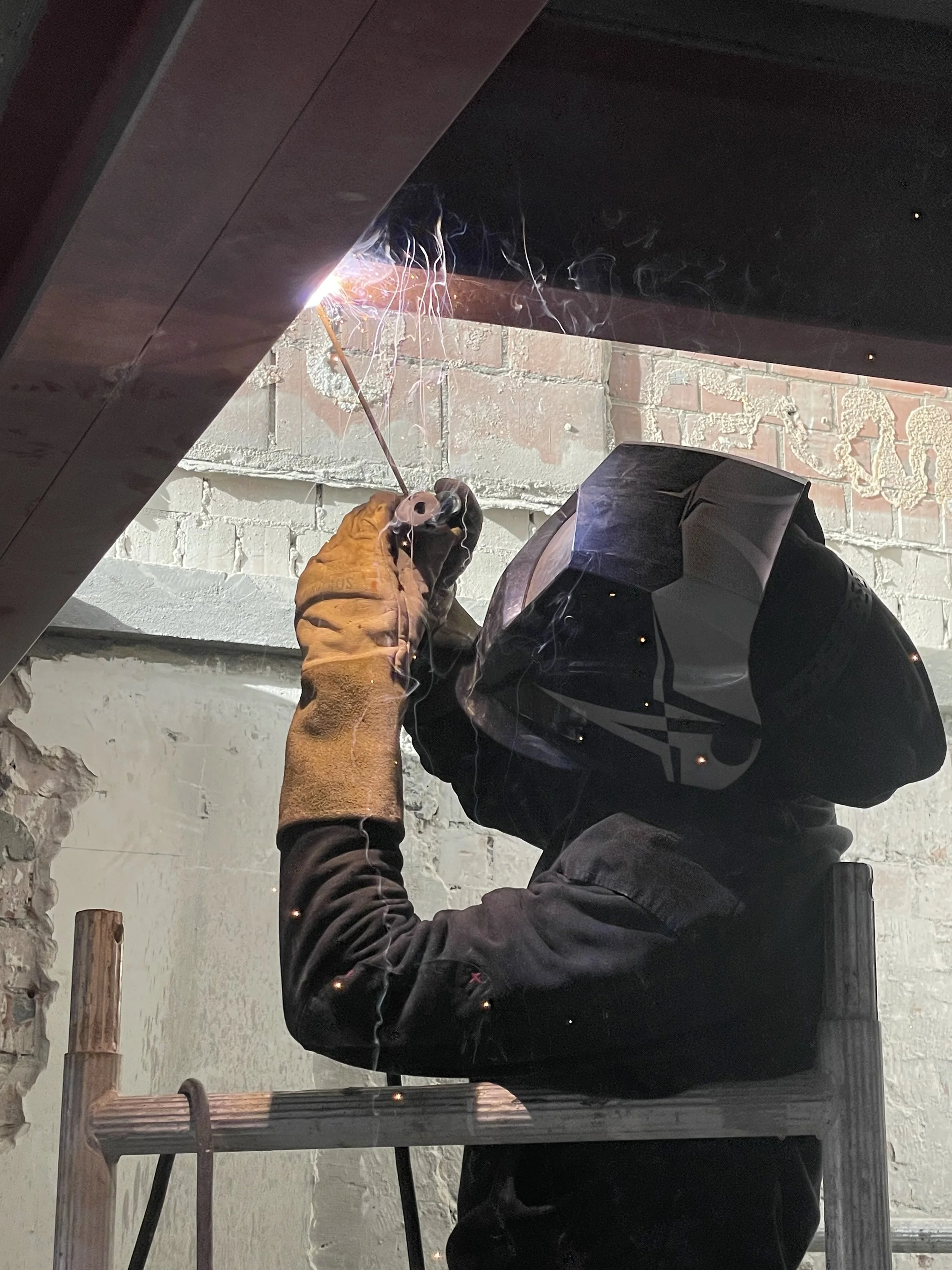 Een persoon die in een werkoutfit en een veiligheidshelm aan het lassen is, met lasvlam en vonken zichtbaar.