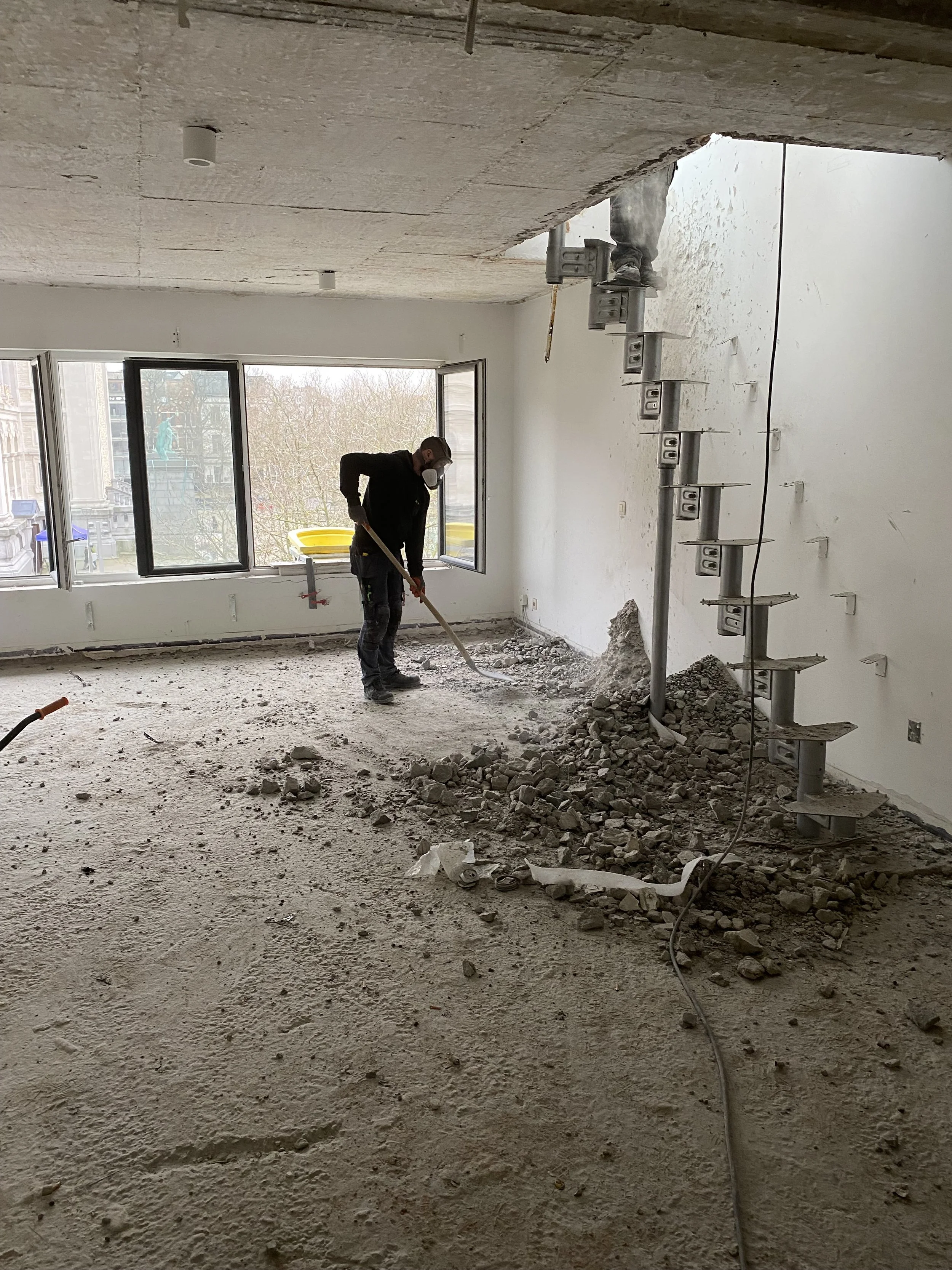 Binnenzicht van een kamer onder renovatie met een man die de vloer aan het egaliseren is. Er is ontmanteld en bouwafval op de vloer, en een deuropening wordt geïnstalleerd.