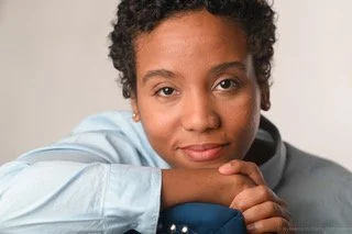 Close-up of a young woman with short curly hair, resting her chin on her hand and looking at the camera
