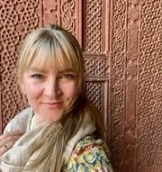 Young woman with blonde hair and bangs smiling outdoors in front of a textured, reddish-brown wall.