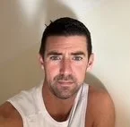 A man with short dark hair and a slight beard, wearing a white tank top, looking into the camera with a neutral expression.