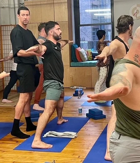 People in a yoga class practicing with blocks and mats, participants in various poses in an indoor studio with wooden floors and large windows.