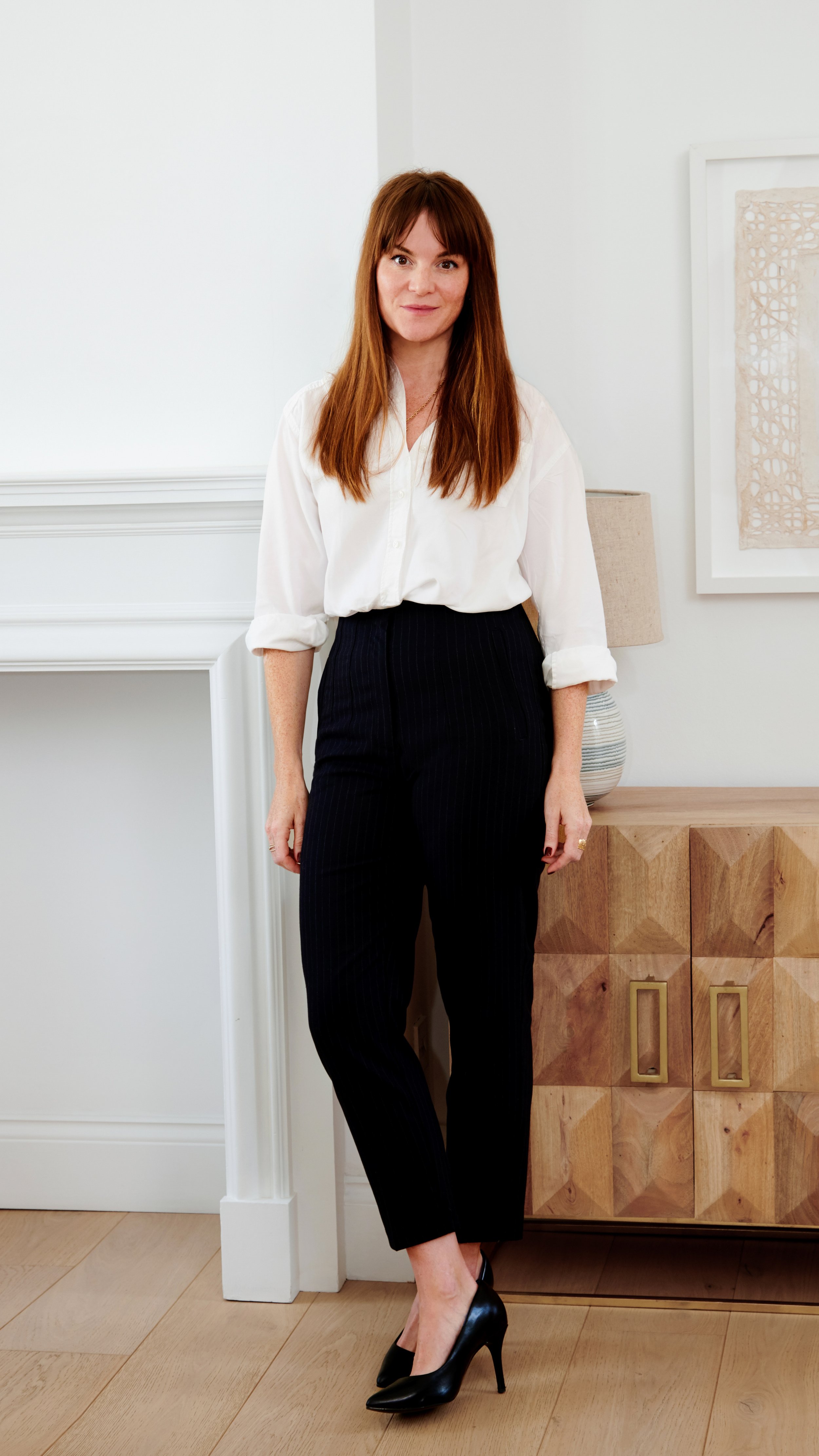 India Wooldridge is wearing a white blouse with sleeves rolled up, black pinstripe pants, and black high heels, standing in a modern, minimalist room with light-colored walls and wooden furniture.