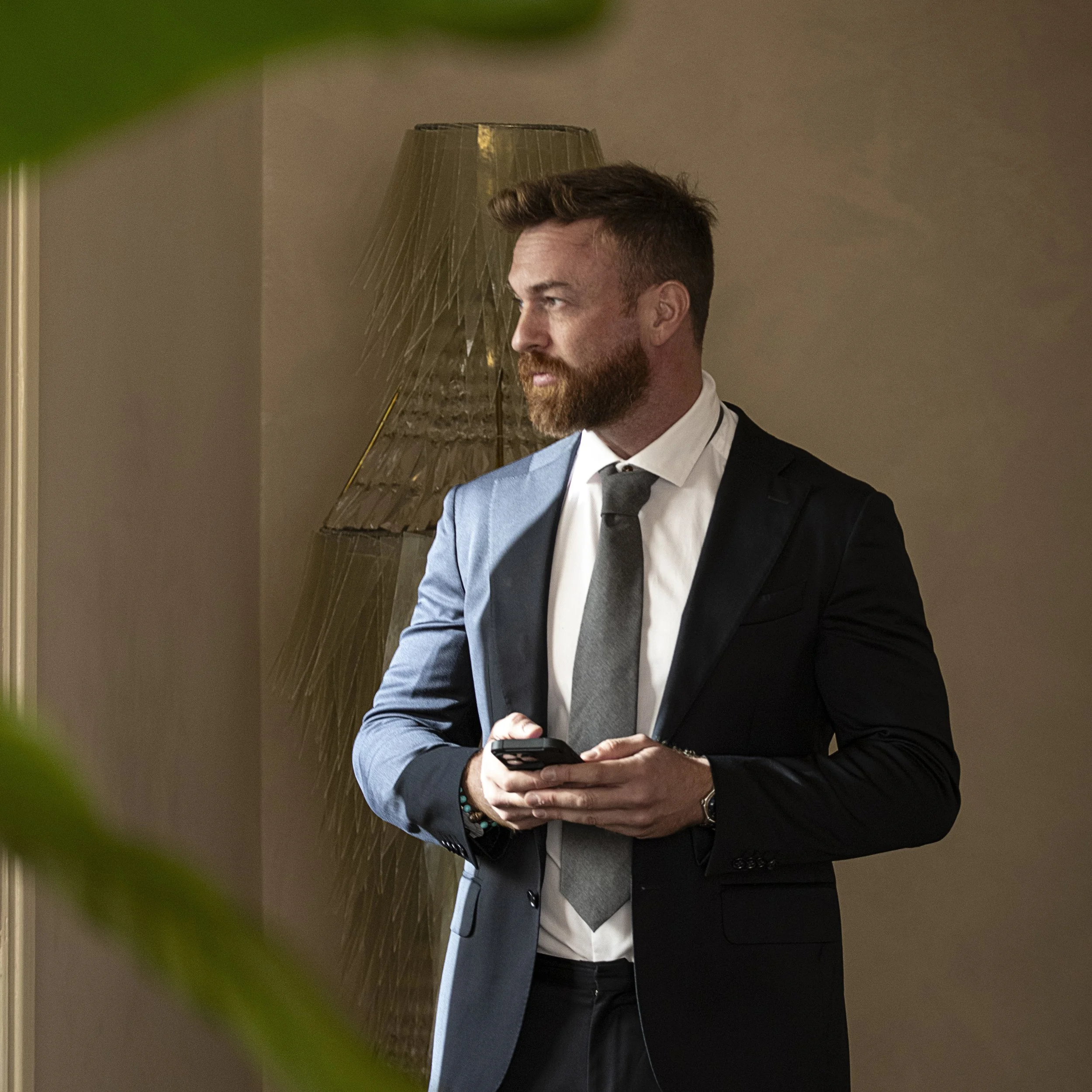 Il fotografo piacentino Andrea Landini in abito elegante nero con cravatta grigia, che tiene in mano uno smartphone, in un ambiente interno con lampada dorata sullo sfondo. Pronto a rispondere alle richieste di preventivi.