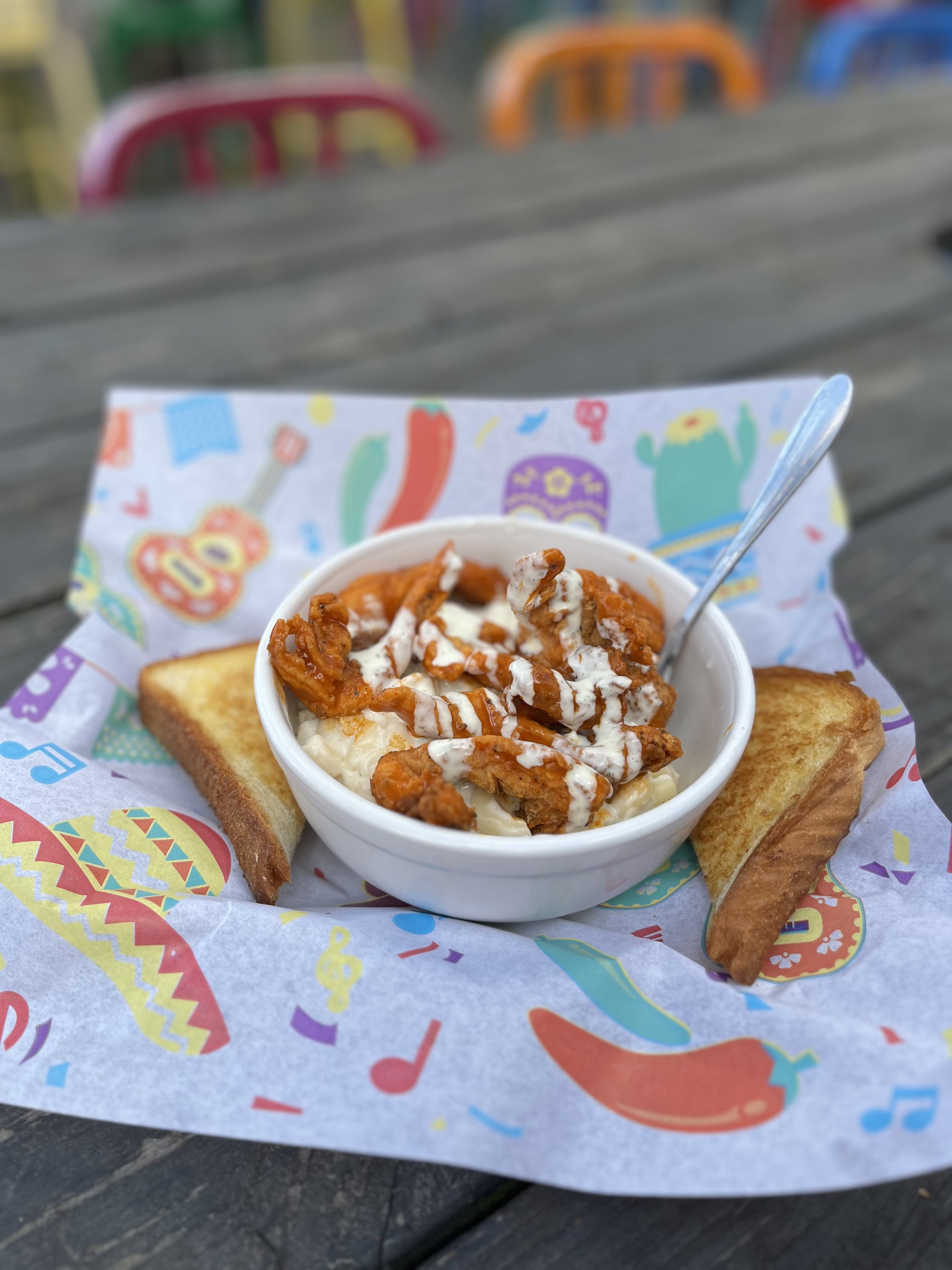 A bowl of mashed potatoes topped with fried chicken strips and gravy, served with two slices of toasted bread on decorated paper at an outdoor setting.