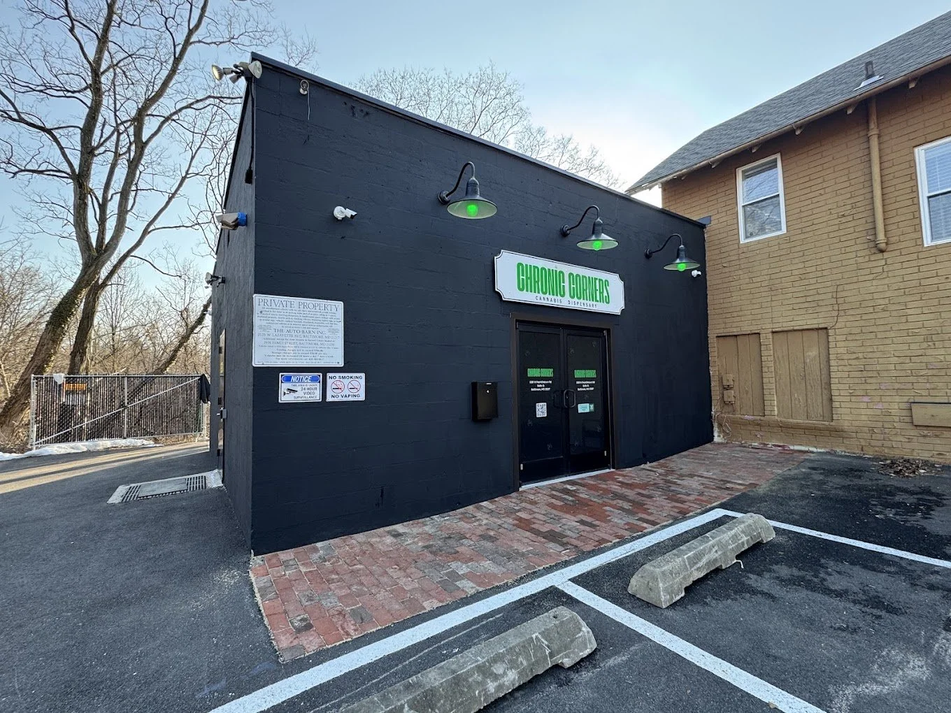 Black building with green sign reading 'Chronic Corners,' located in a parking lot with brick and asphalt paving, next to a beige brick building, with leafless trees in the background.