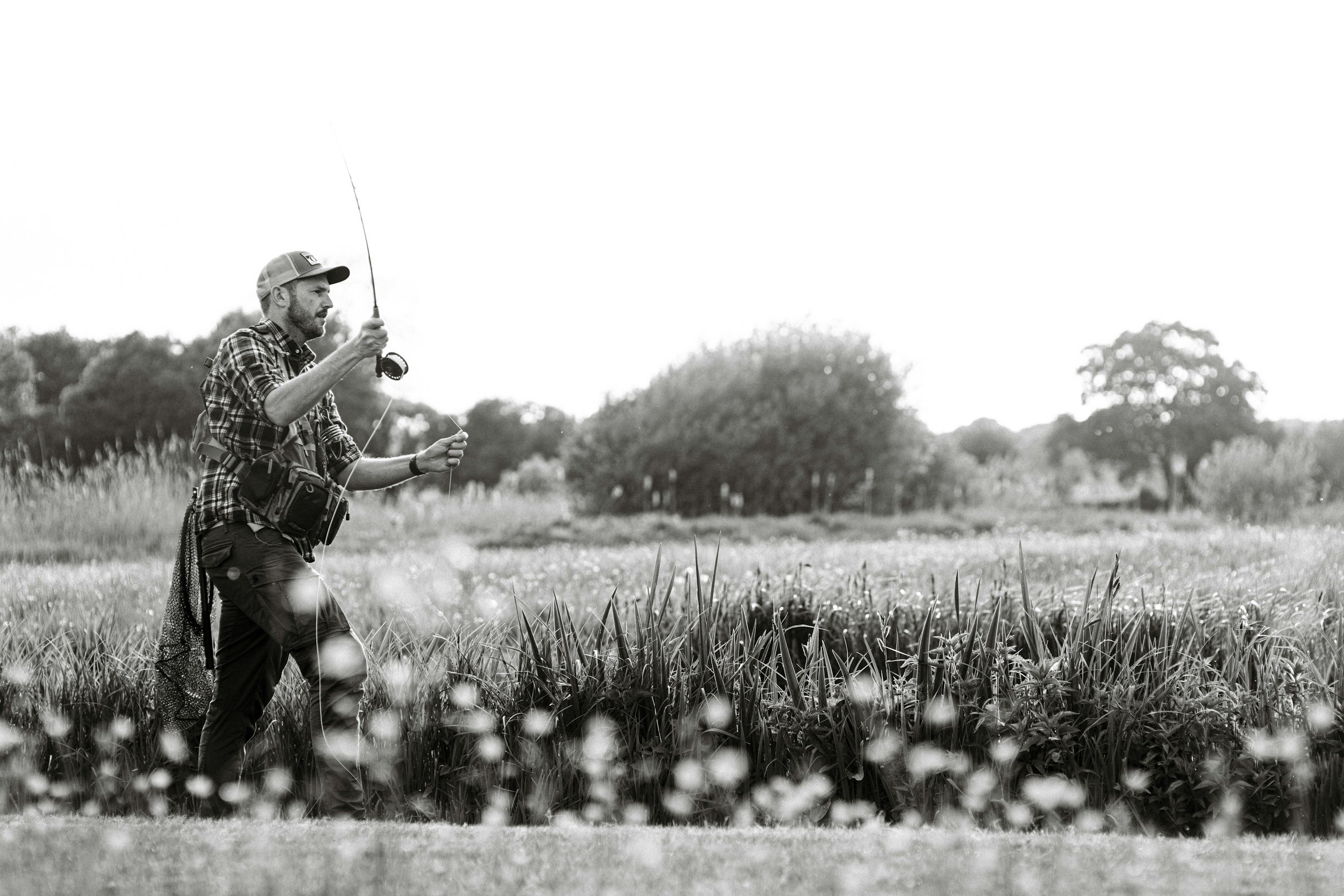 Black and white image of a person fly fishing in a field with tall grasses, wearing a plaid shirt and cap, surrounded by trees.